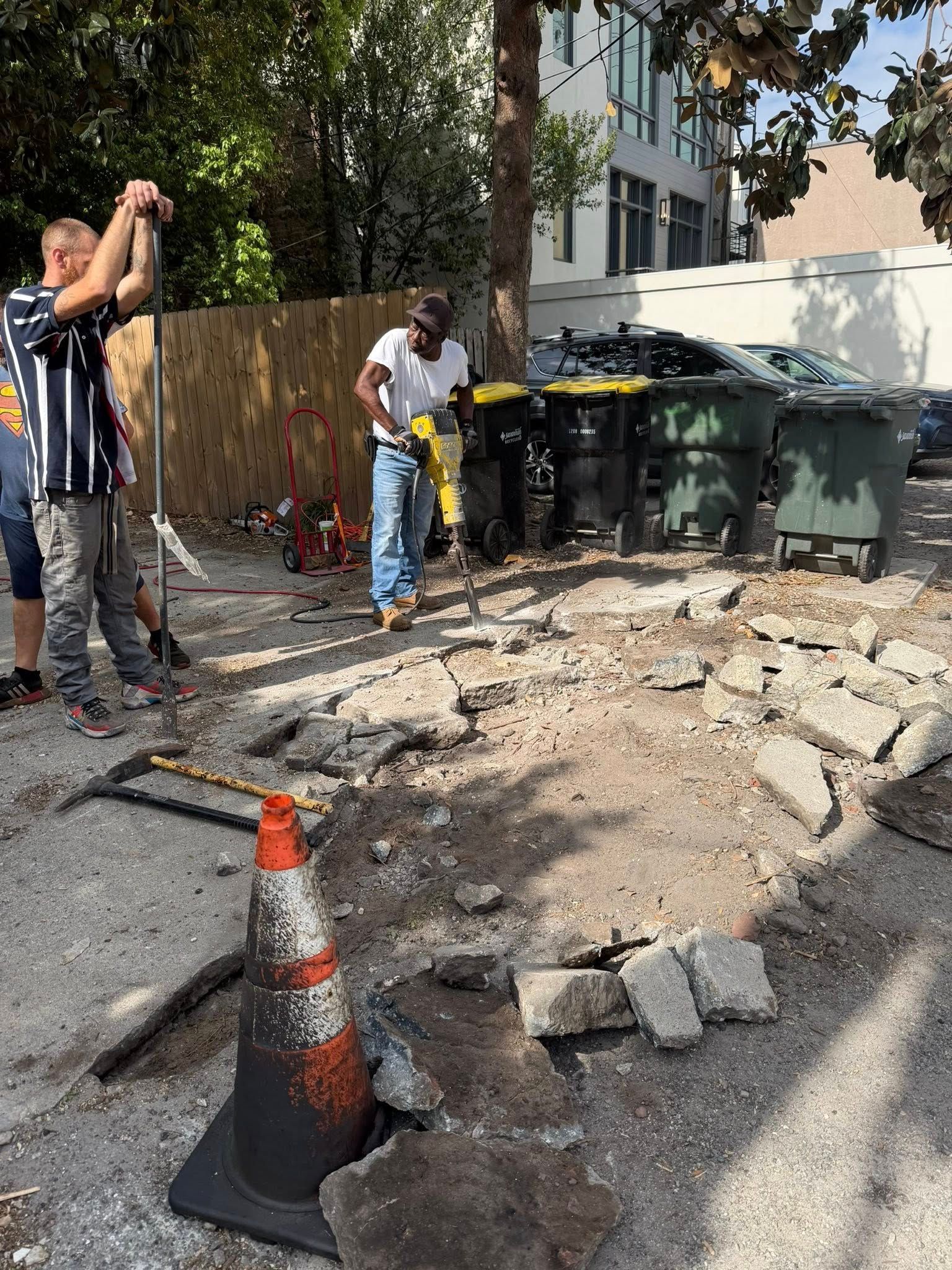 Workers repairing a broken sidewalk beside trash bins, with rubble and traffic cones in the foreground.