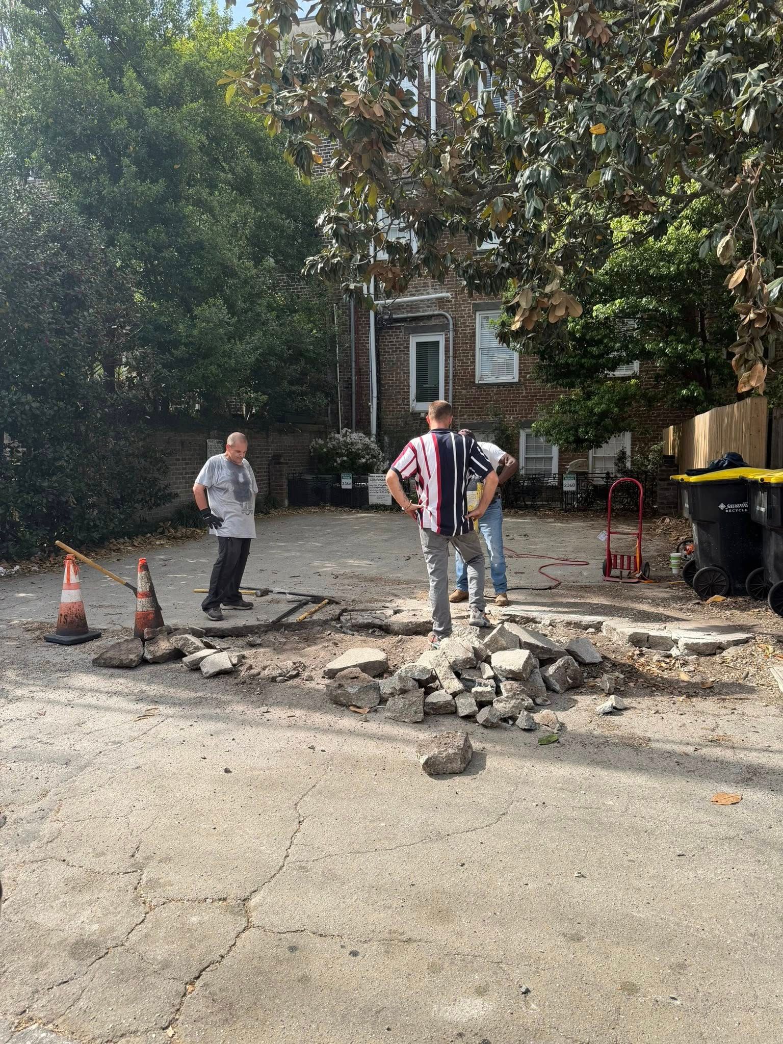 Workers and a stroller near broken pavement and orange cones on a tree-lined street