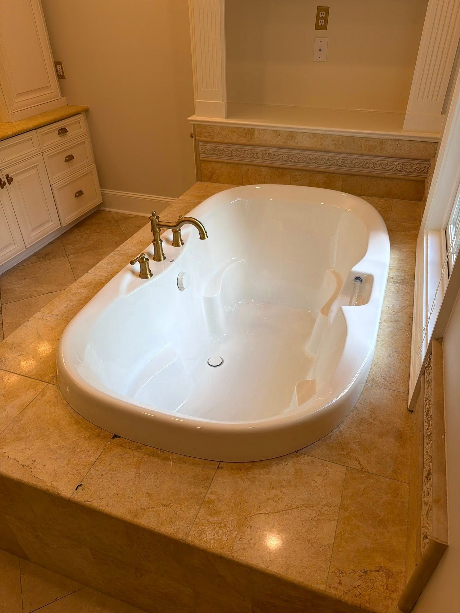 Cream-colored bathroom with a sunken white soaking tub and beige stone surround.