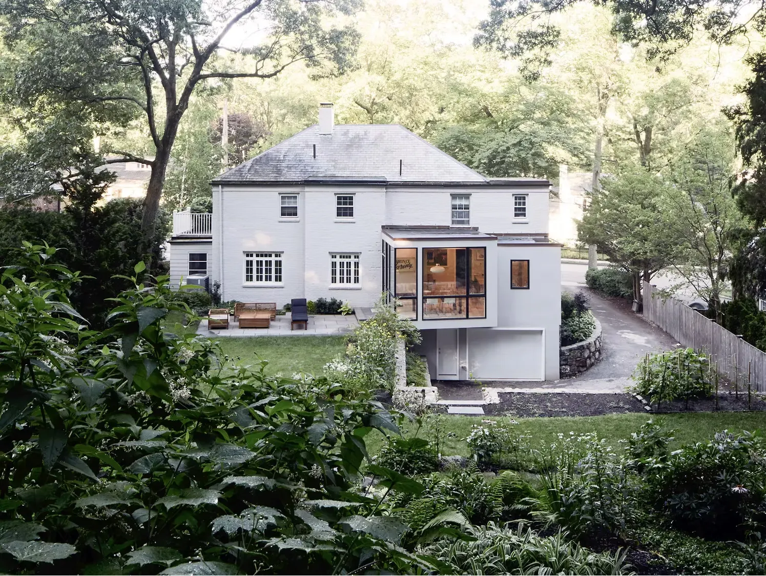 White two-story house with a modern glass extension, surrounded by lush green trees and garden.