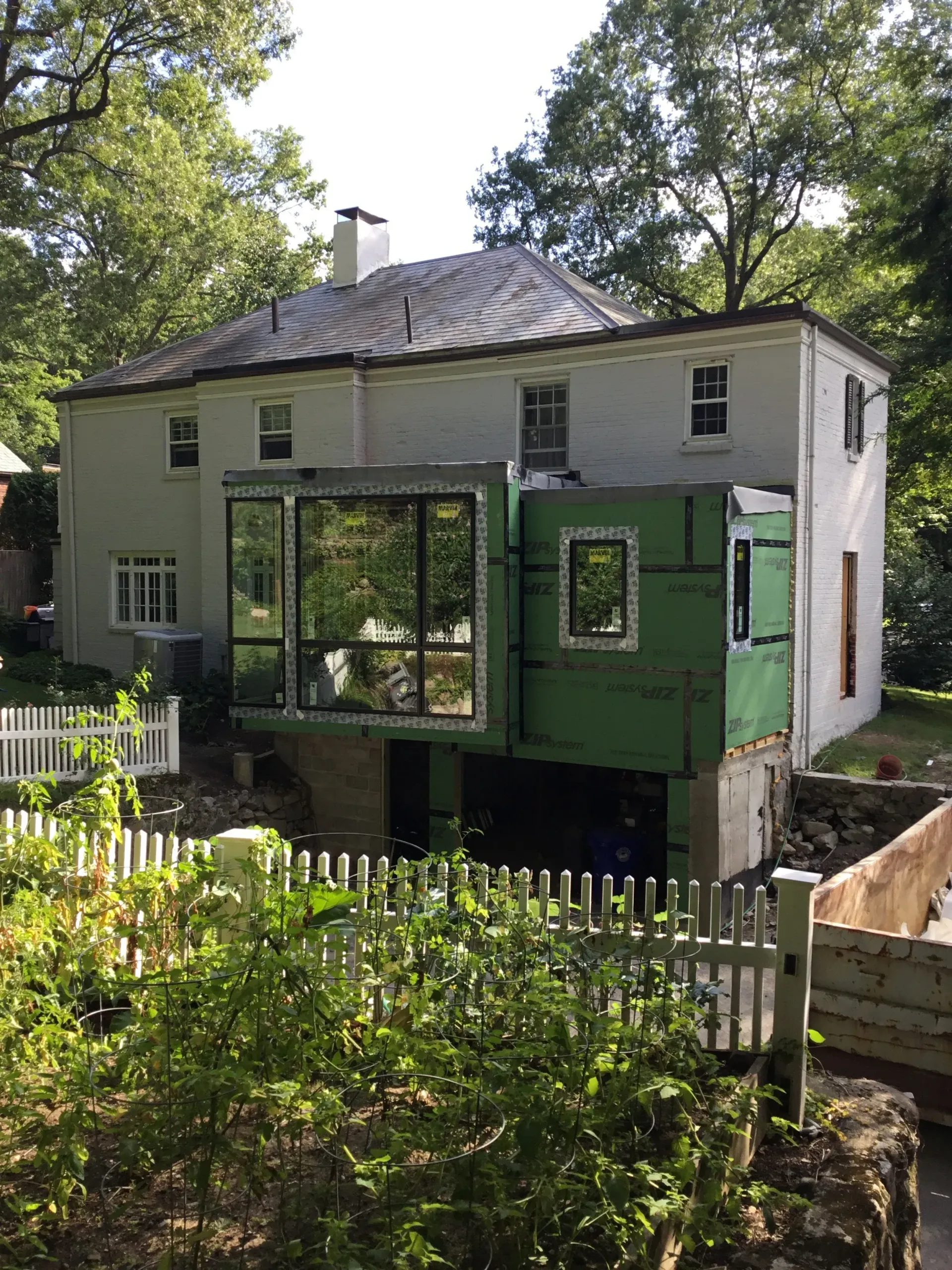 White house with dark roof and green porch addition behind a picket fence in a leafy yard