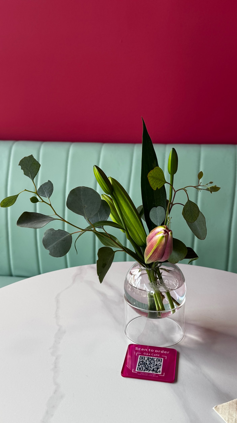 Small glass vase with flowers on a white table, against a mint-green bench and pink wall.