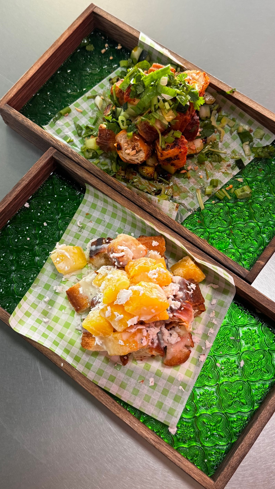 Two trays of glazed bite-sized dishes on checkered paper in a wooden serving box, garnished with herbs and oranges