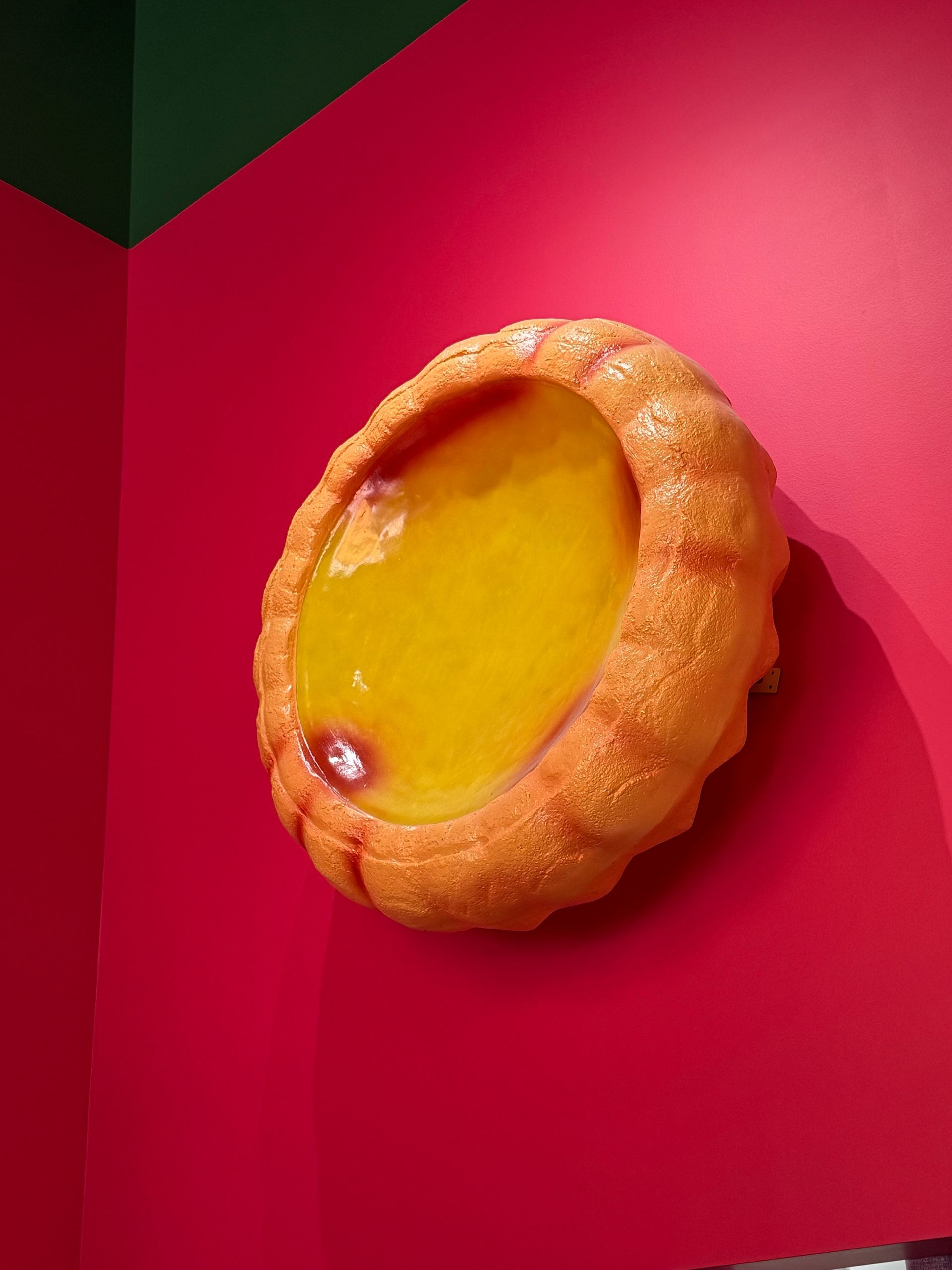 Yellow tart-like pastry mounted on a bright pink wall, viewed at an angle.