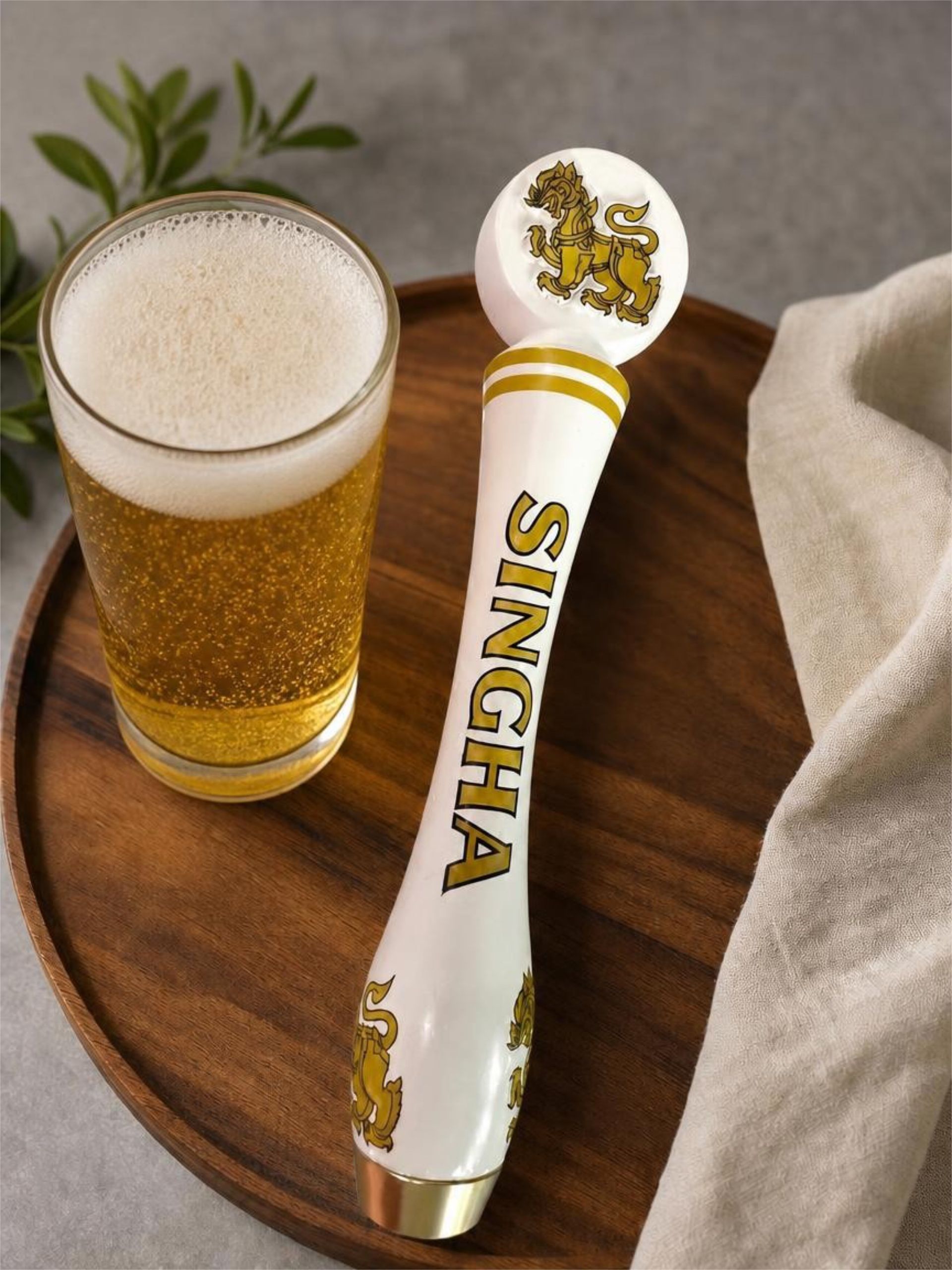 A glass of beer beside a white and gold Singha-branded bottle opener on a gray fabric surface