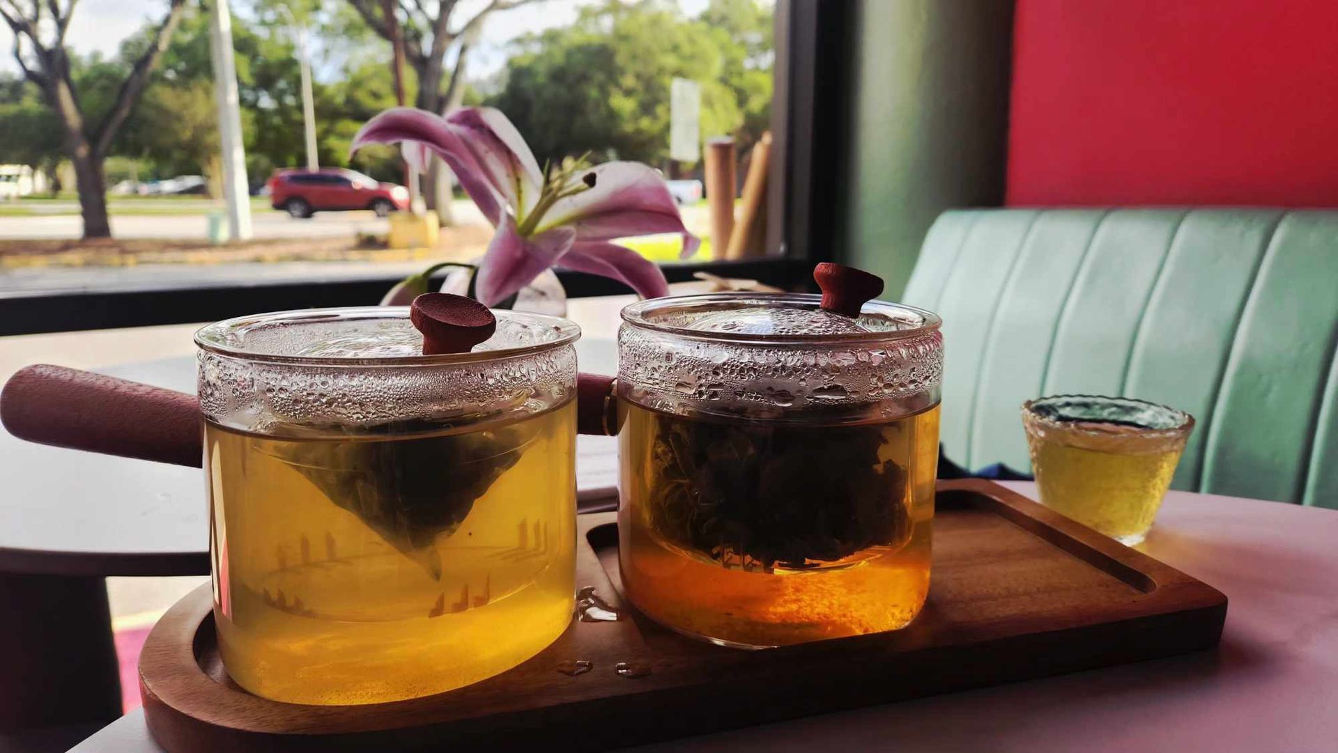 Two iced drinks on a wooden tray by a cafe window, with a small flower garnish and a mint-green seat behind.