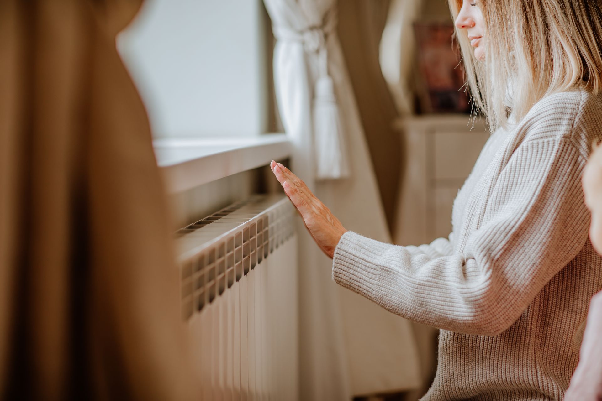 A woman in a long sweater is holding her hands near the radiator at home.