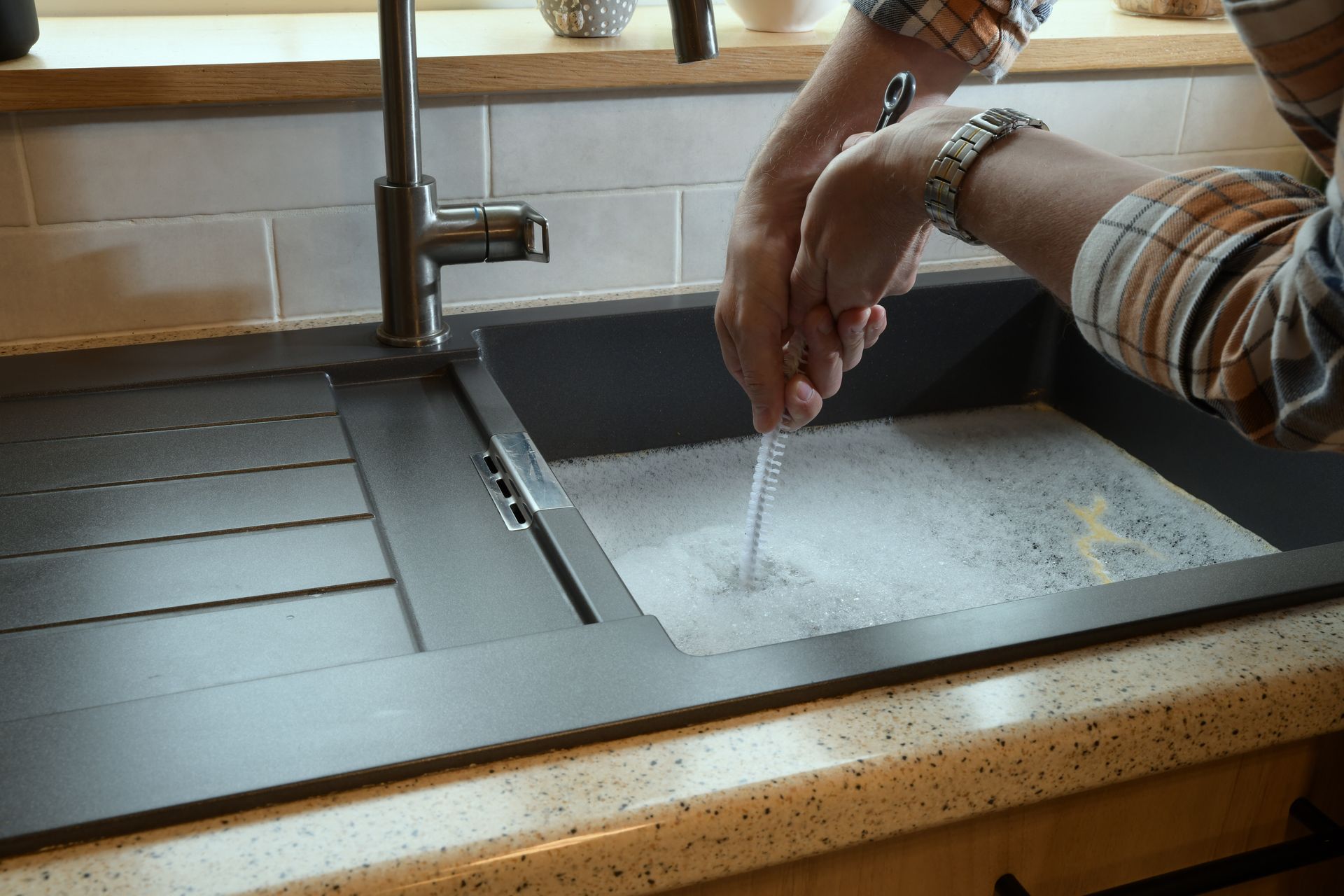 Person uses a drain snake tool to clear a clogged kitchen sink filled with soapy water.