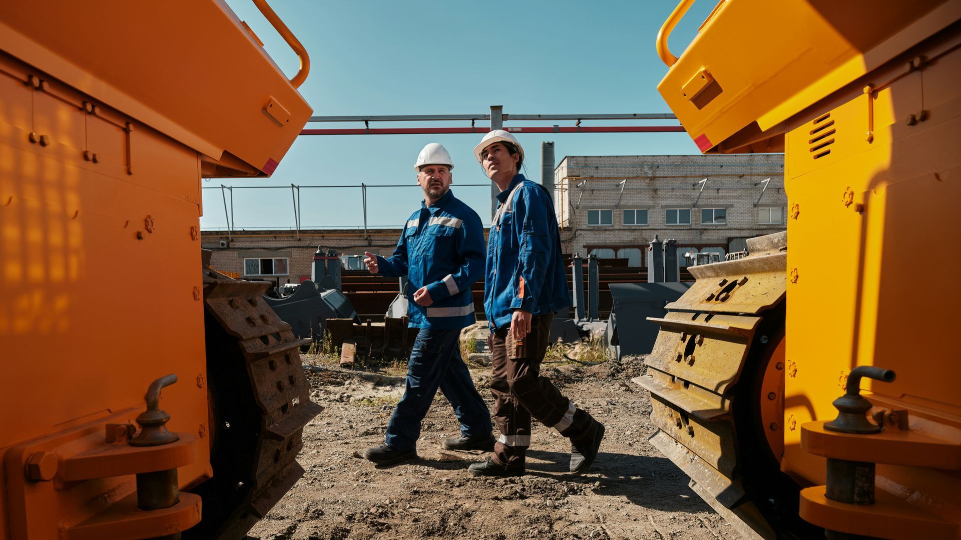 Two workers in hard hats stand between large yellow construction machines on a dirt site