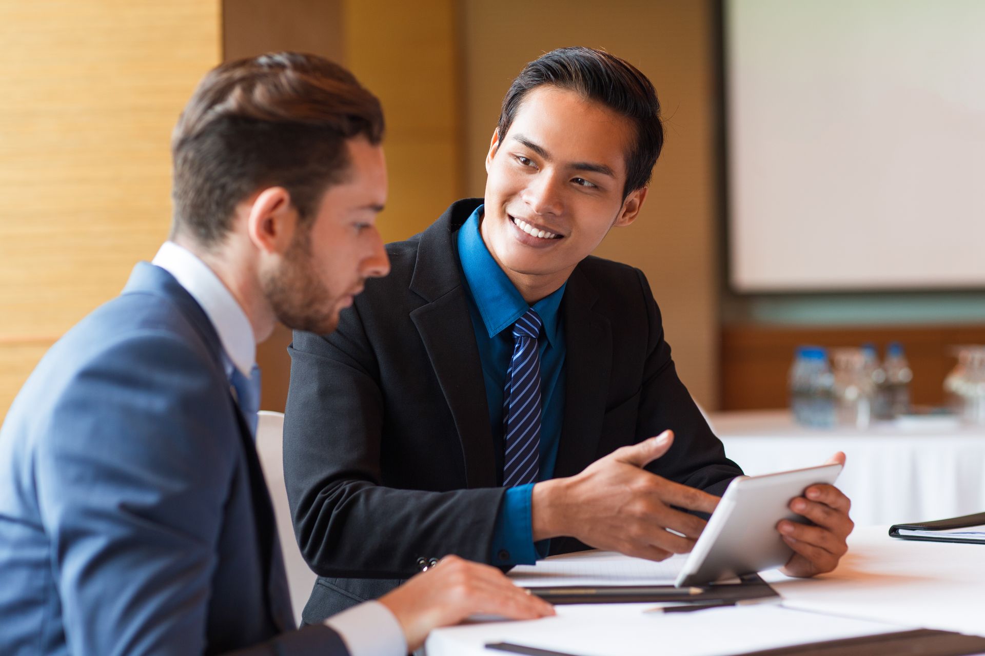Two professionals in a meeting, one holding a tablet and smiling across a conference table