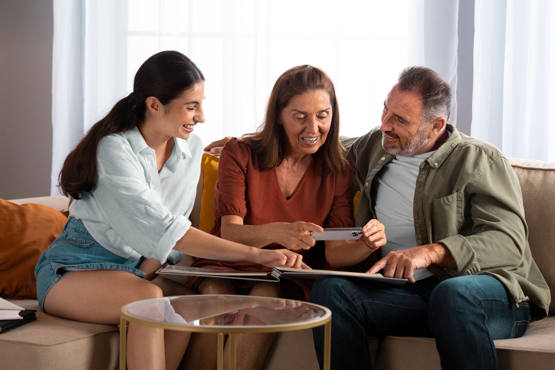 Three people sit on a couch smiling as they look at a tablet together in a living room.
