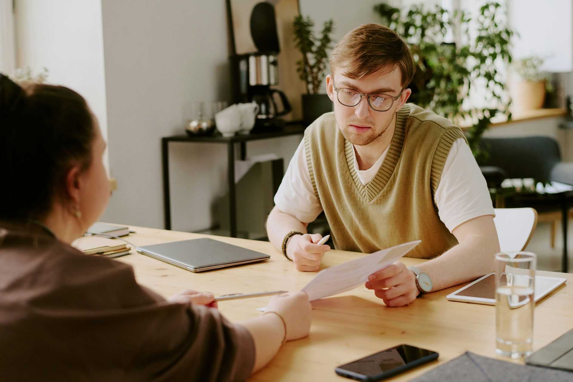 Two people discussing papers at a desk in a bright office, with a laptop, phone, and water glass nearby.