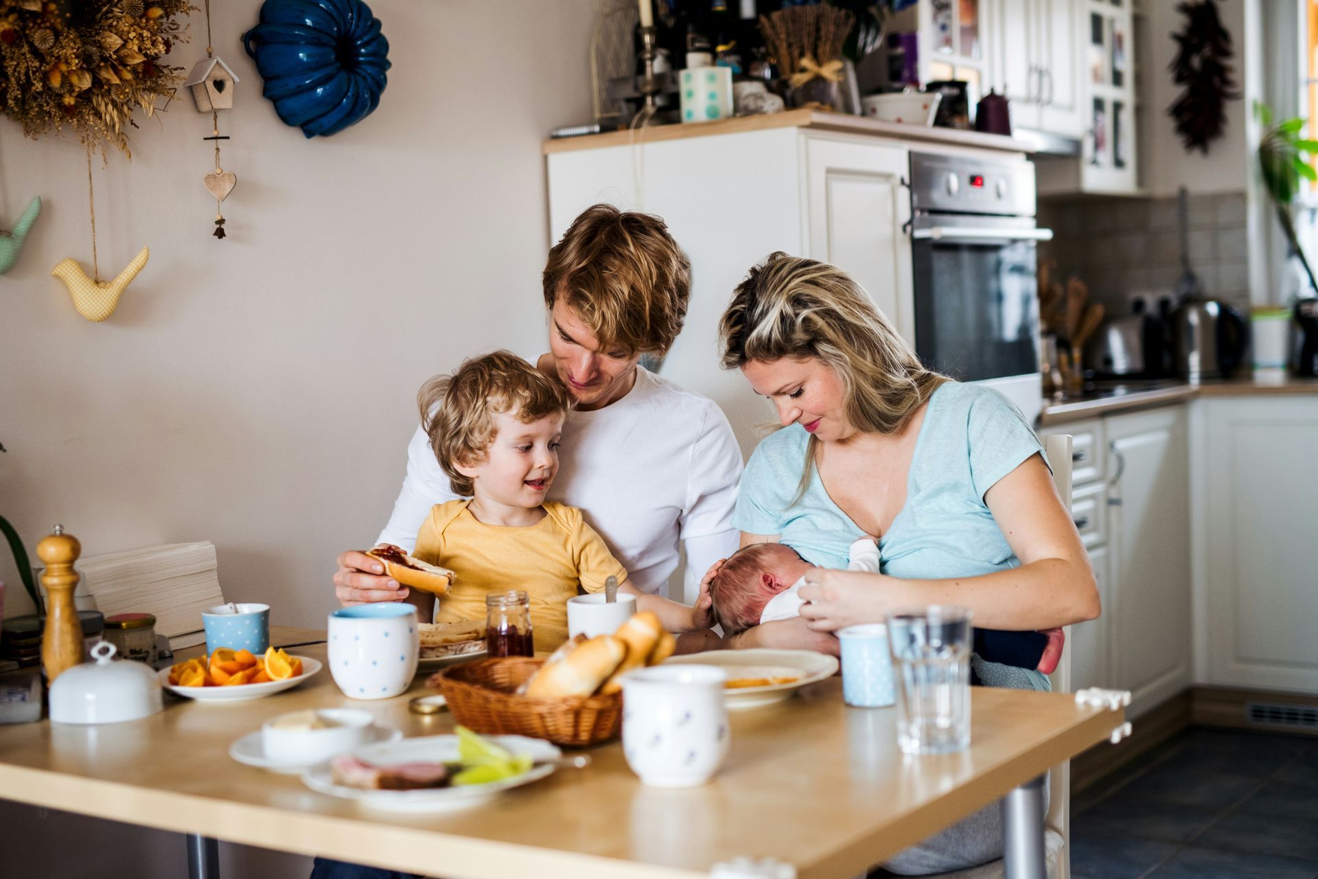 Family baking together at a kitchen table, smiling and mixing ingredients with bread and mugs nearby