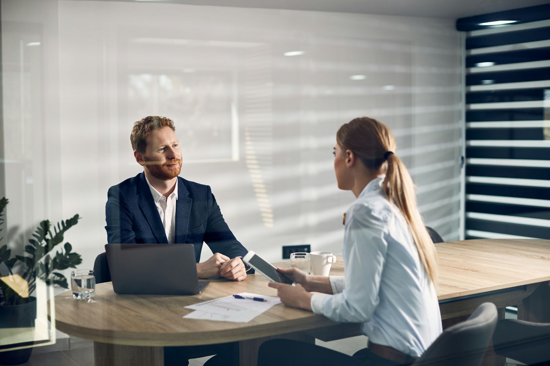 Two coworkers talking across a conference table in a bright office, with a laptop and notebook open.