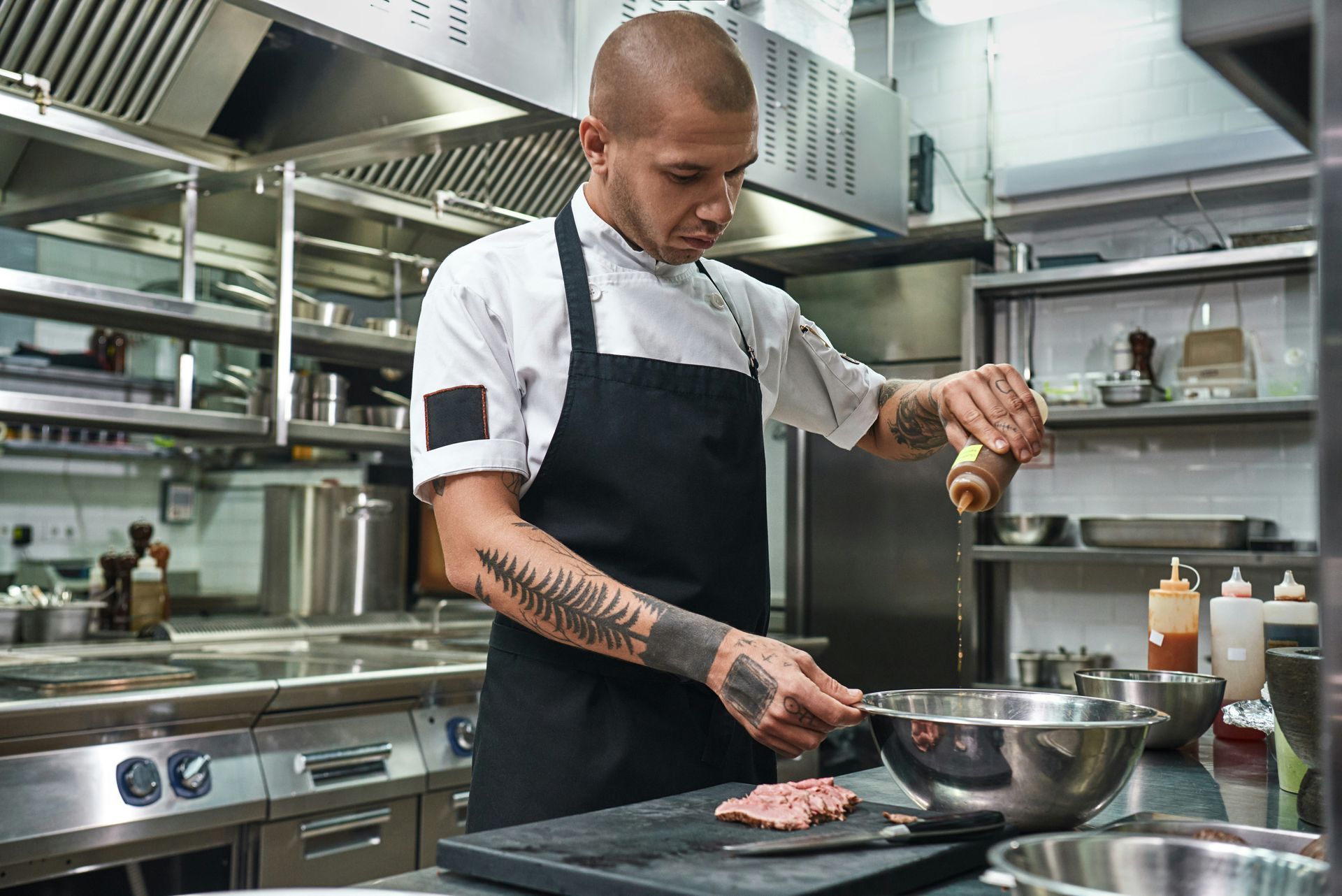 Chef seasoning raw meat in a stainless-steel kitchen, with cookware and prep tools nearby.