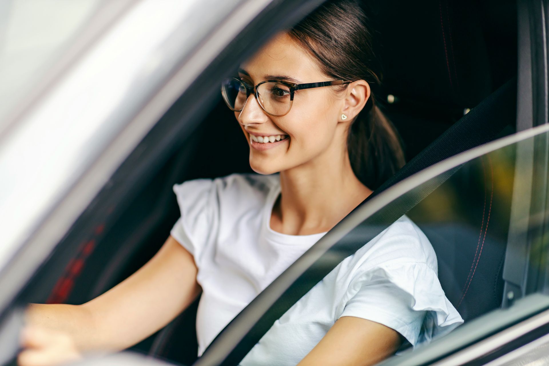 Smiling woman in glasses driving a car, seen through the window.