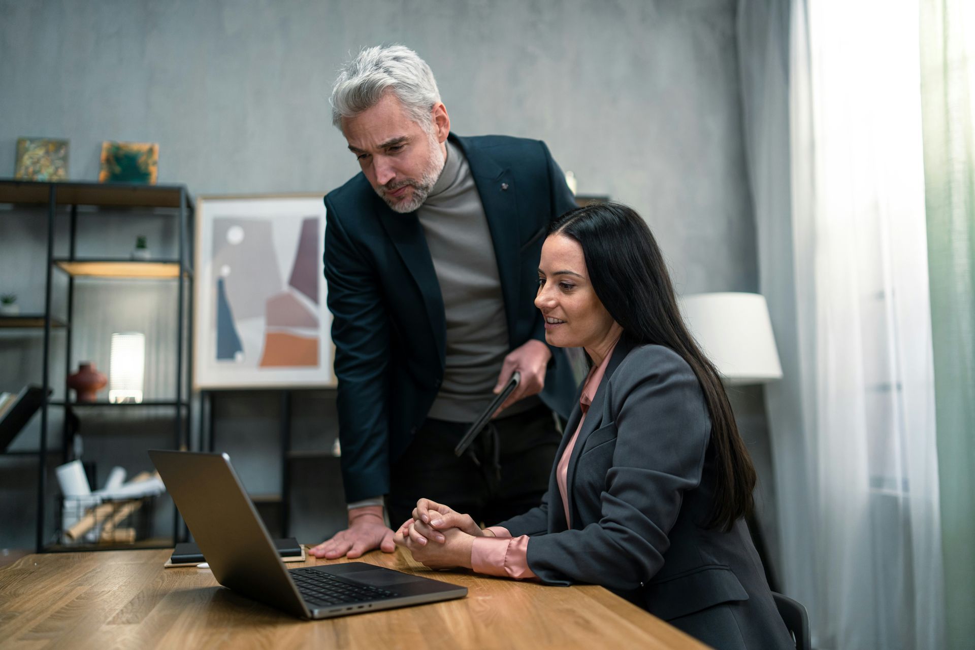 Two coworkers reviewing a laptop at a desk in a modern office, one standing and leaning over the other.