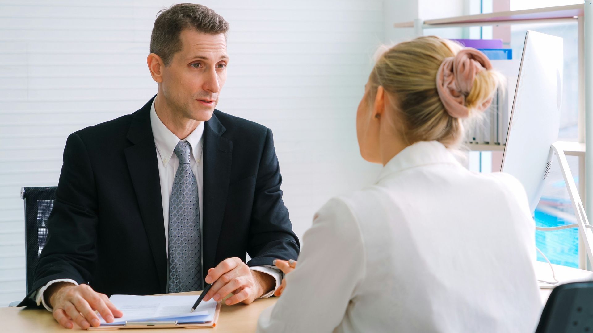 Two people in a business meeting across a desk, discussing documents in a bright office.