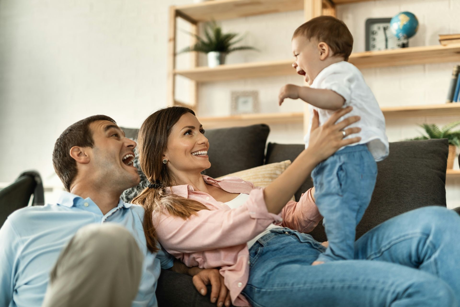 Family sitting on a couch, smiling as a child is lifted by an adult in a bright living room.