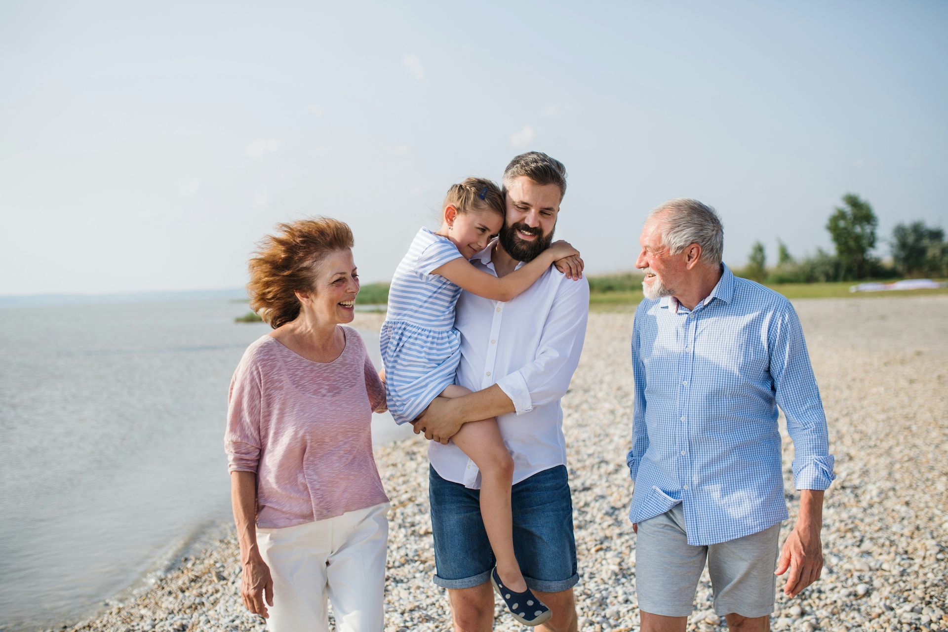 Four people walking together on a rocky beach by the water, smiling in the sunlight.