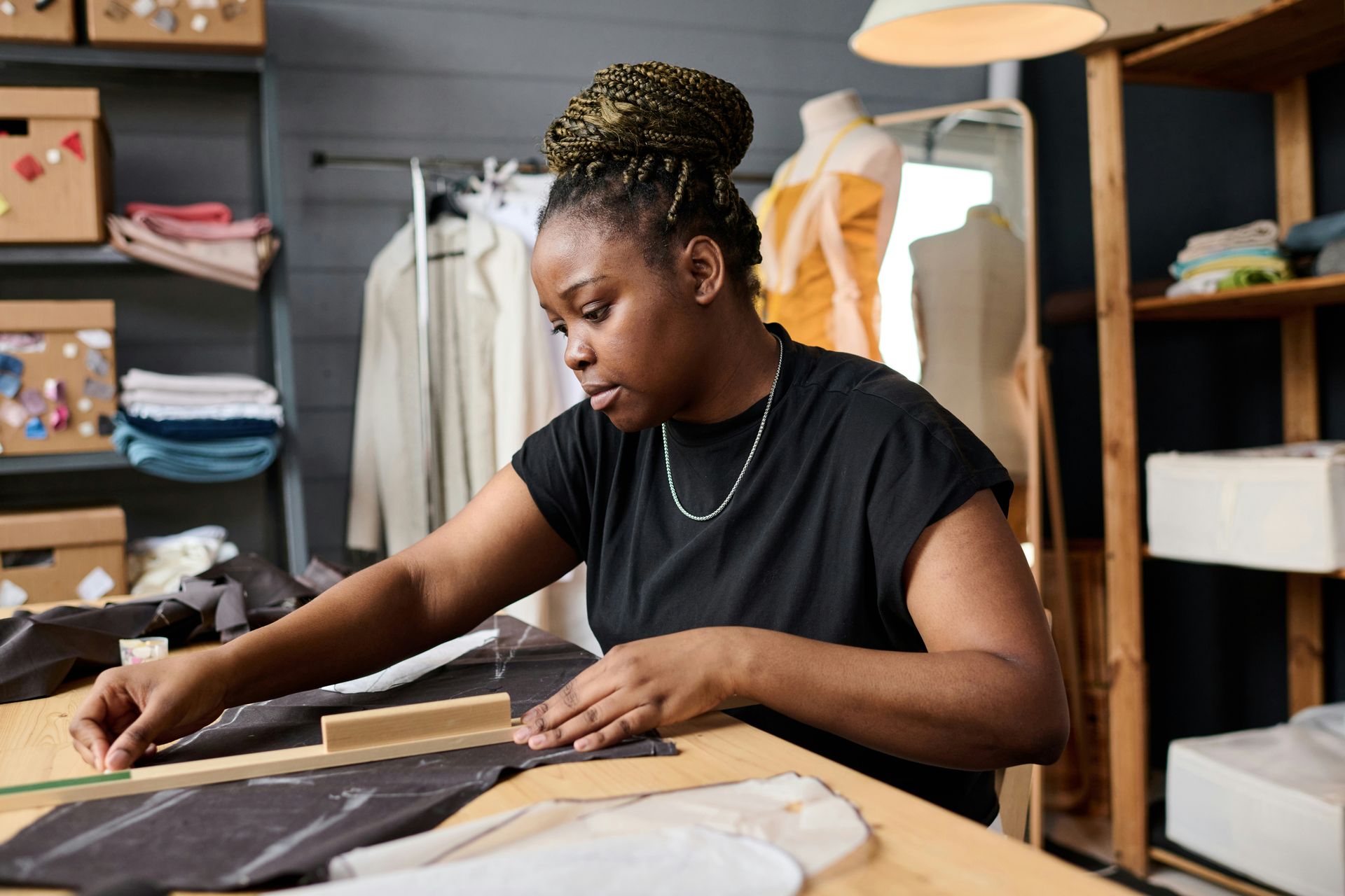 Person measuring fabric at a sewing table in a workshop with shelves and hanging clothes