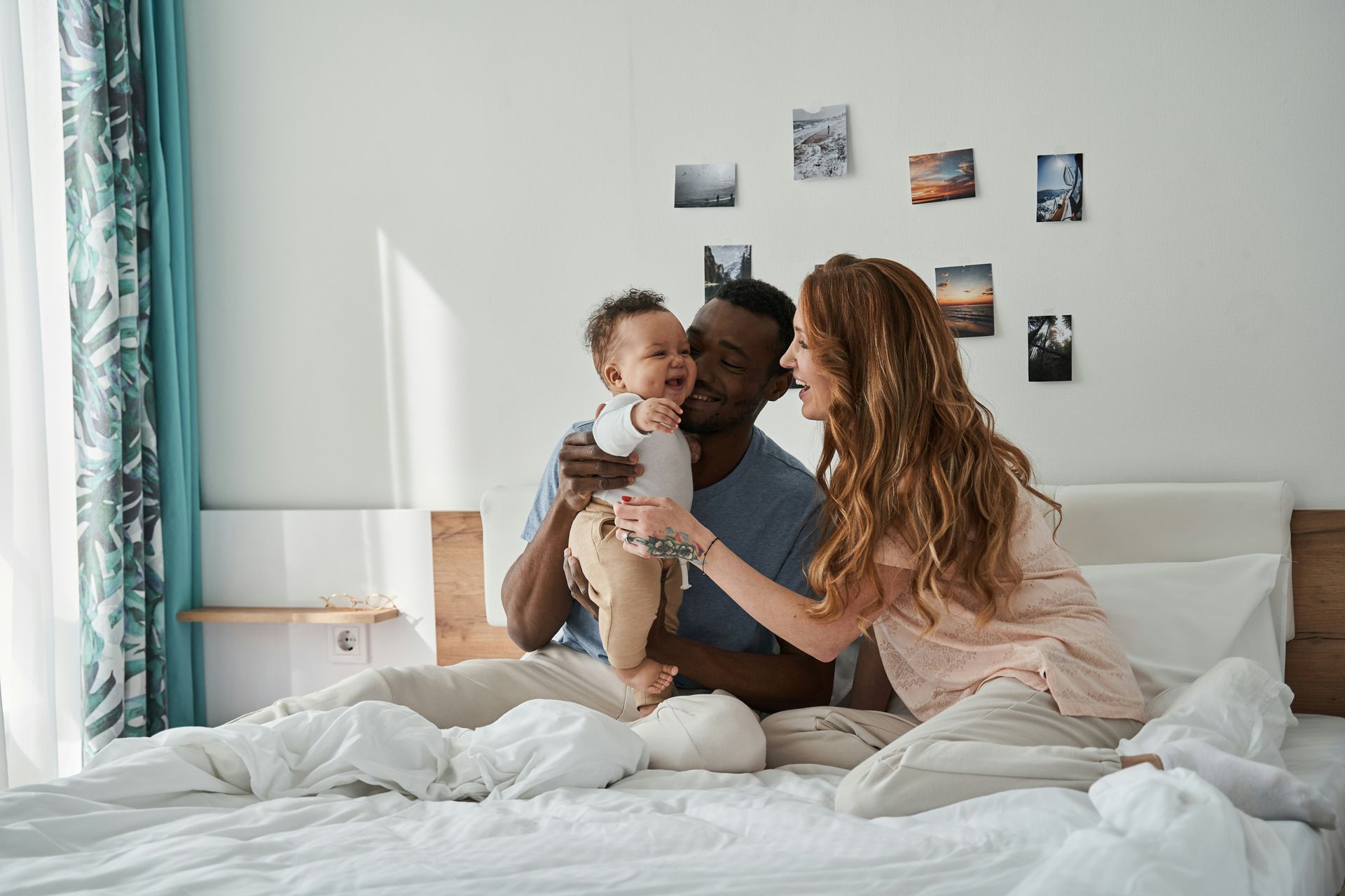 Family sitting on a white bed, holding a baby in a bright bedroom