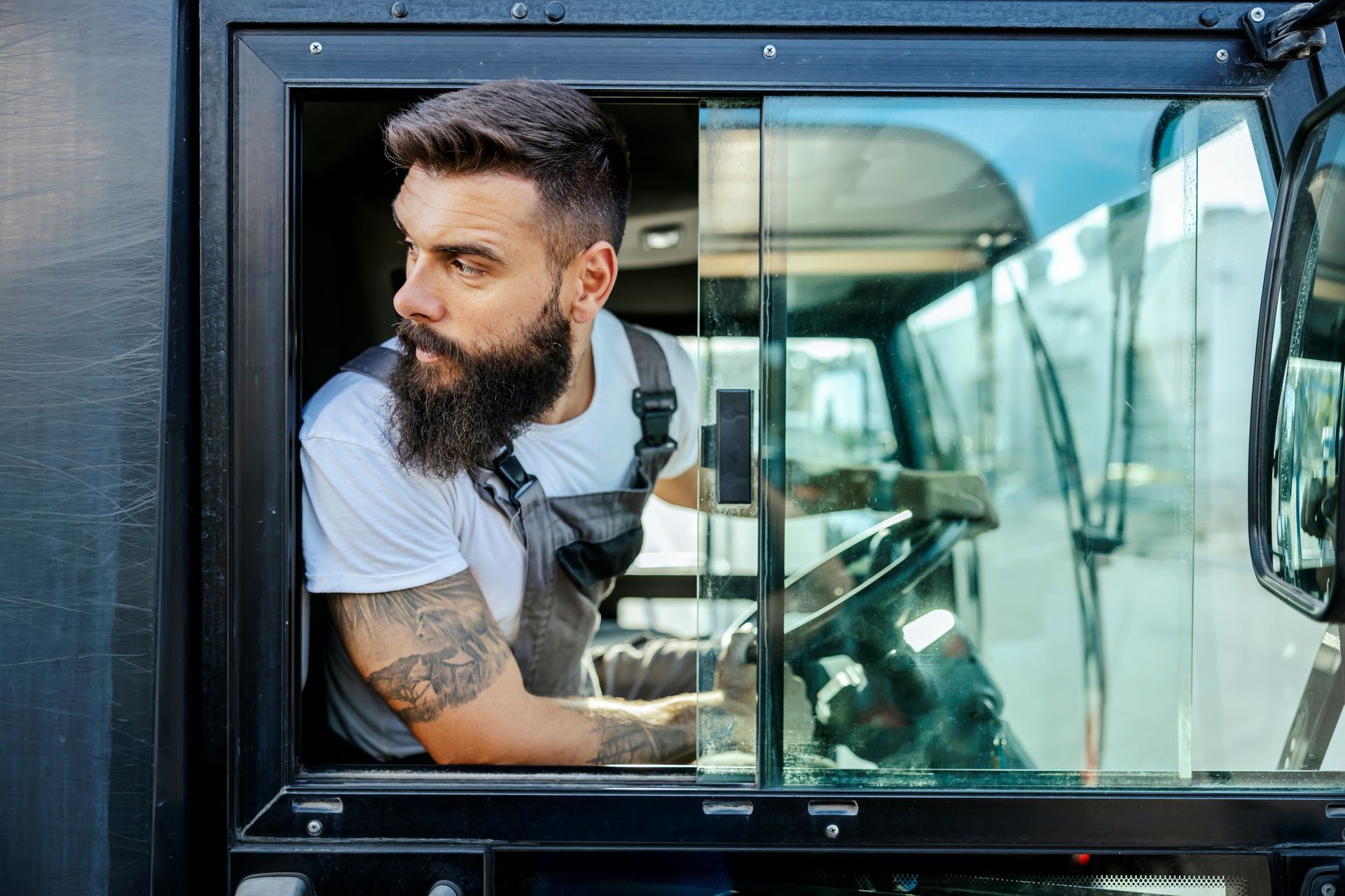 Bearded man in a white shirt driving a vehicle, seen through the open window.