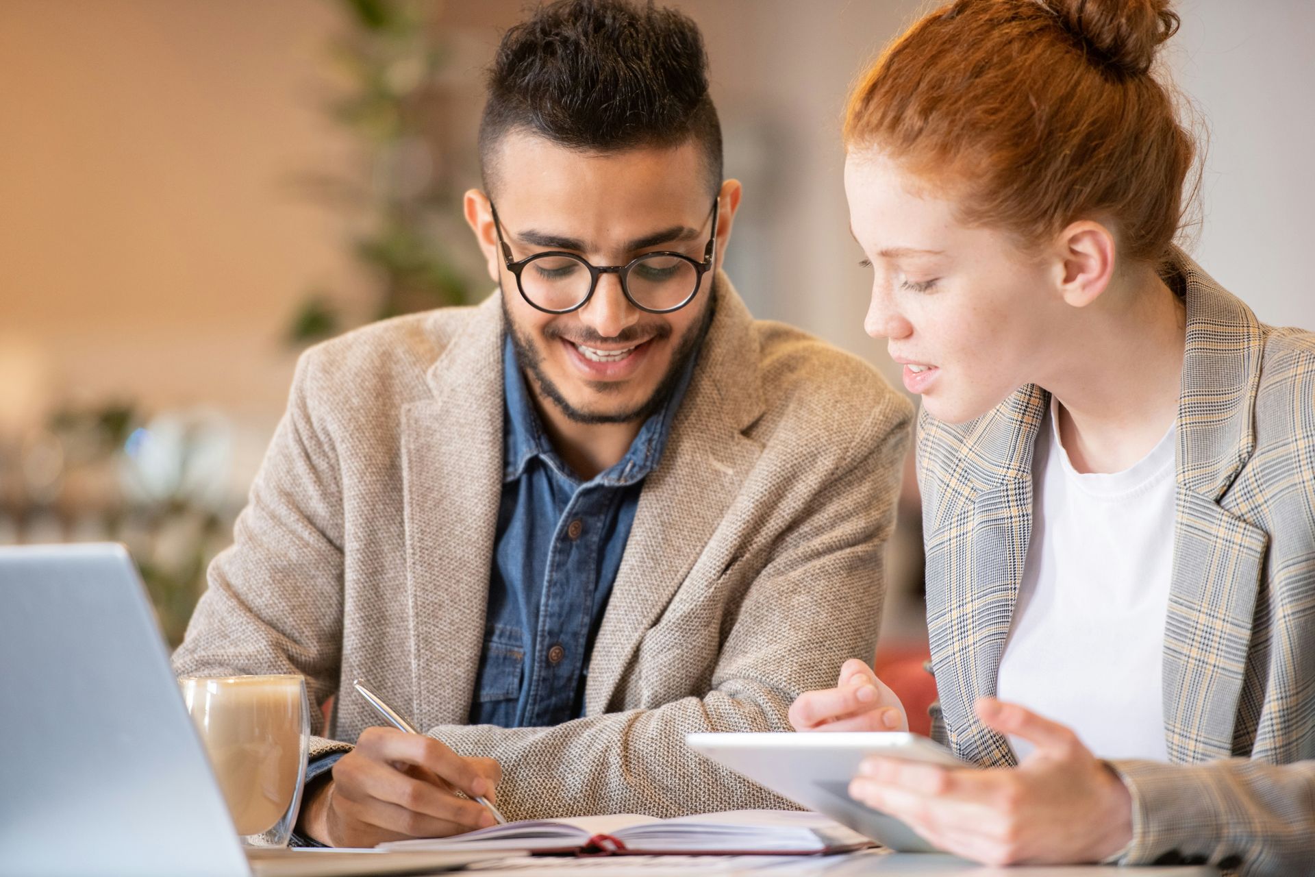 Two coworkers smiling and reviewing papers at a table with a laptop and coffee