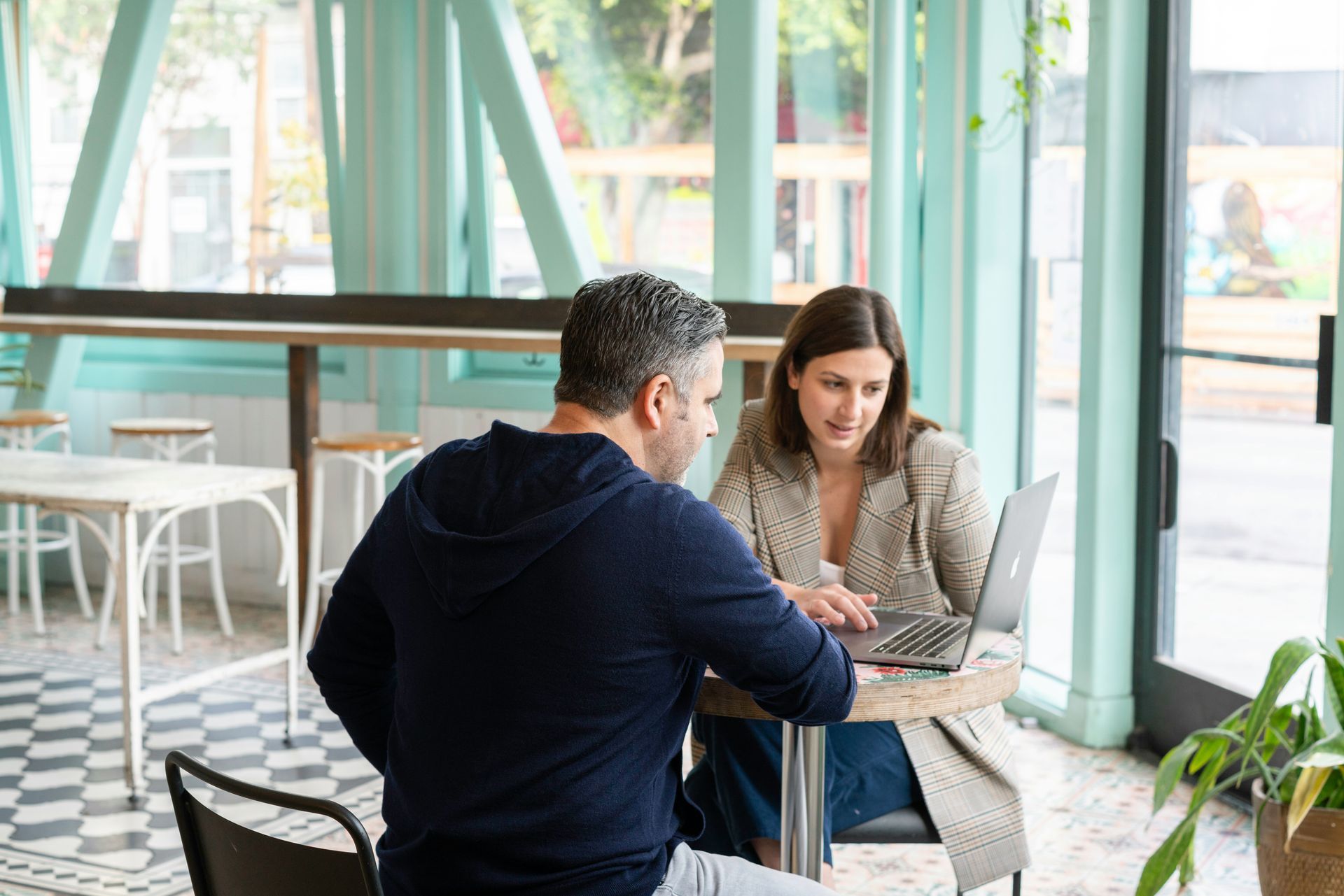 Two people discuss a laptop at a café table near large windows