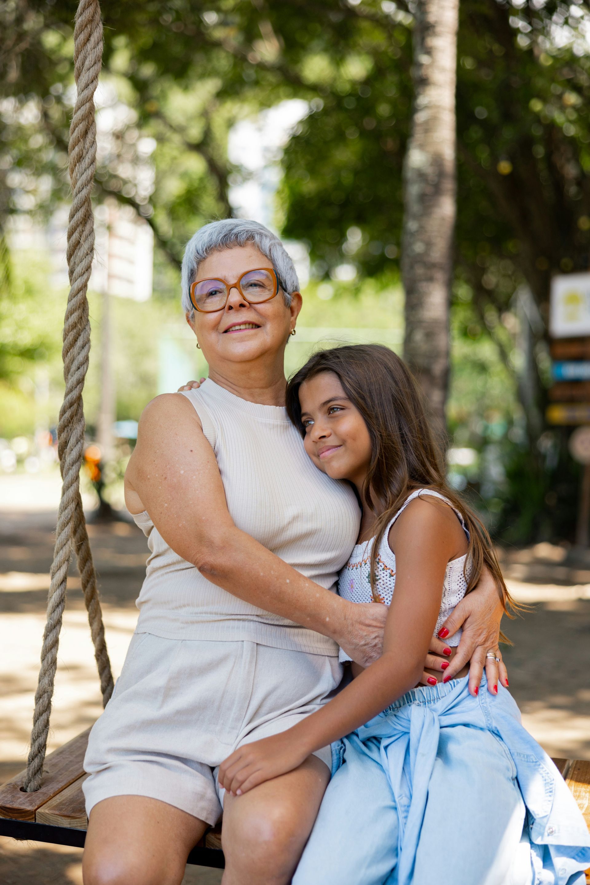 Older woman and young girl smiling on a park bench, with trees and greenery in the background.