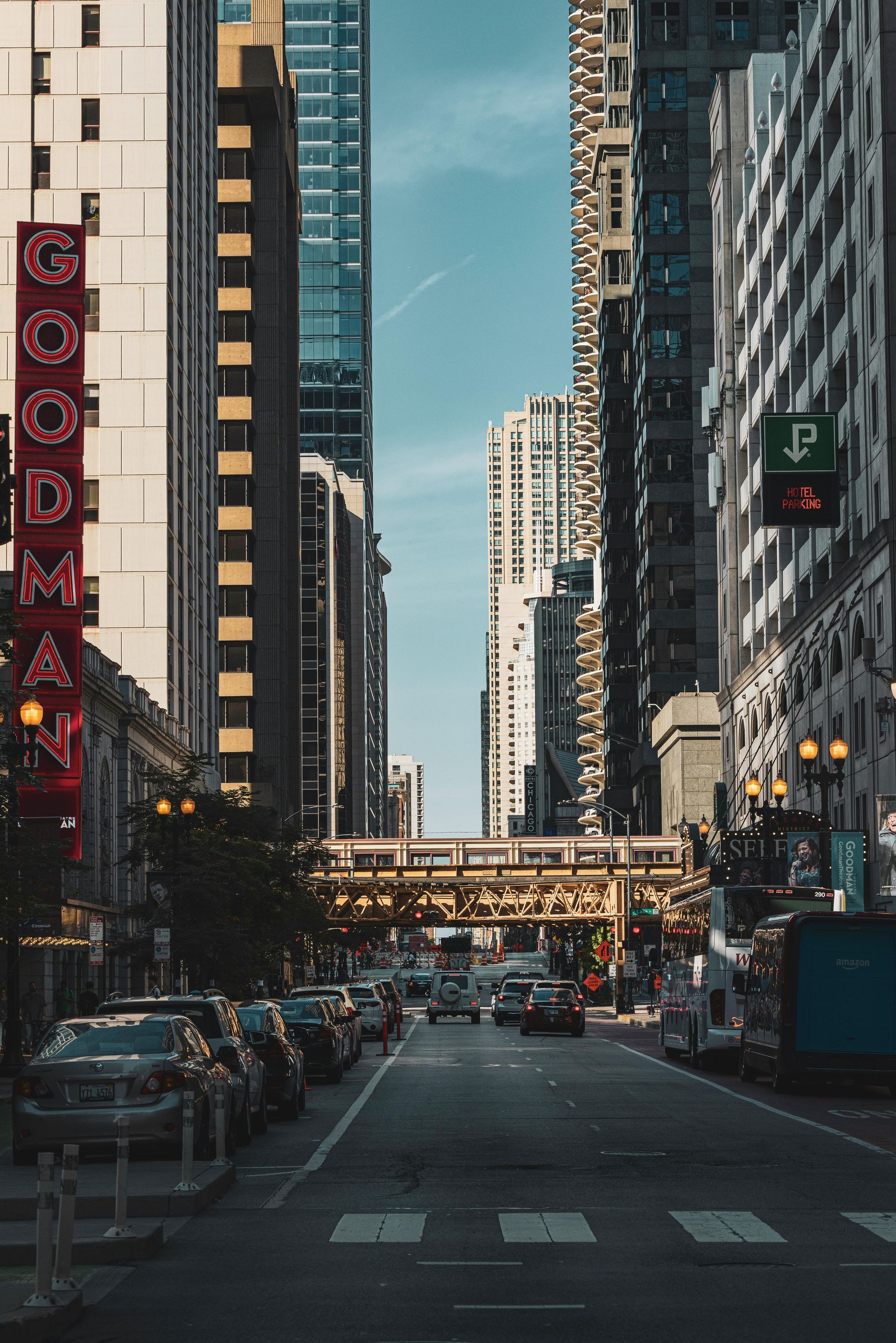 City street canyon with tall buildings, cars, and a pedestrian bridge under a clear blue sky