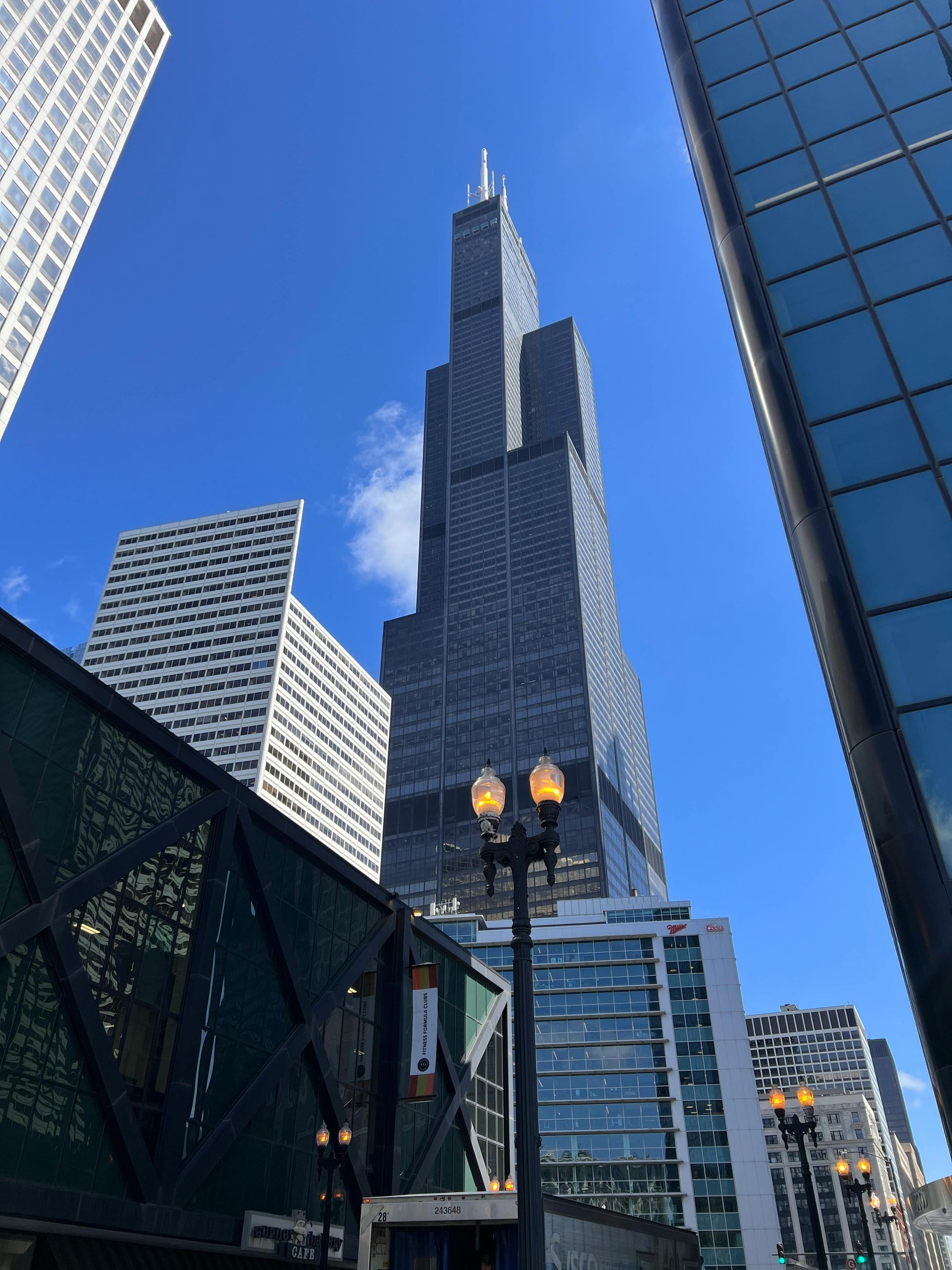 Tall glass skyscraper against a blue sky, framed by surrounding buildings in an urban cityscape.