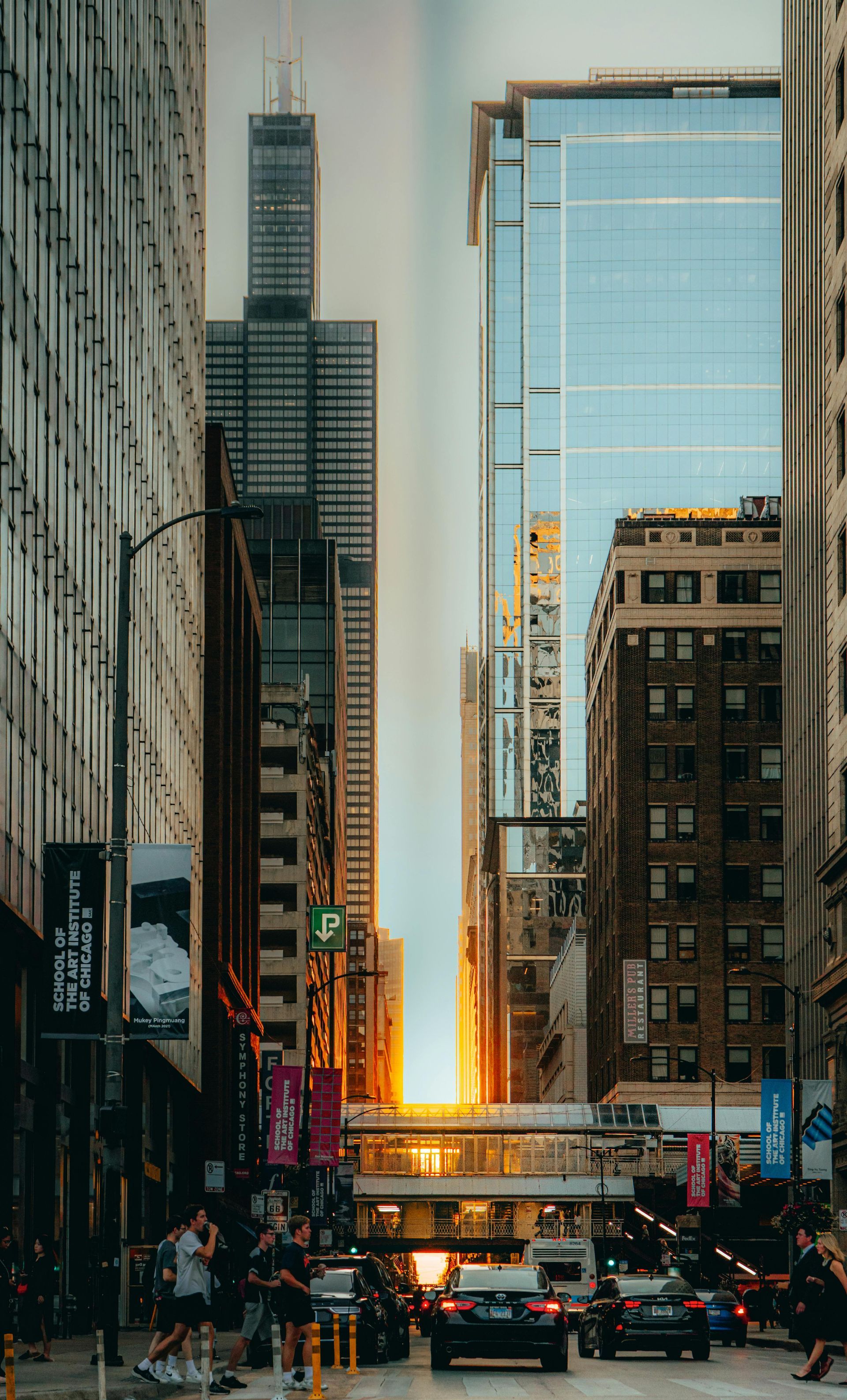 Downtown city street between tall buildings, with traffic and a glowing sunset at the end of the avenue