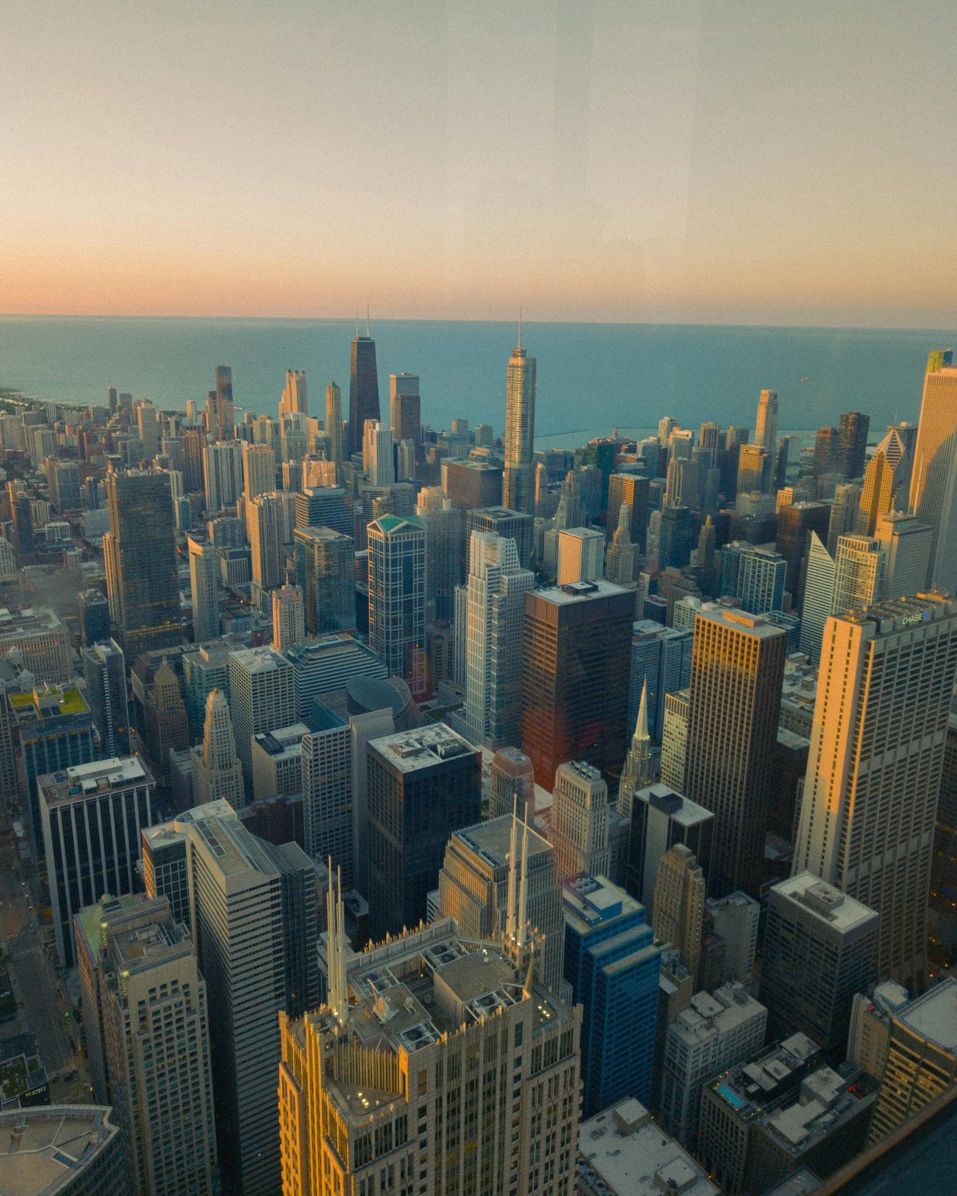 Aerial view of a dense city skyline by the water at sunset, with tall skyscrapers in warm light.
