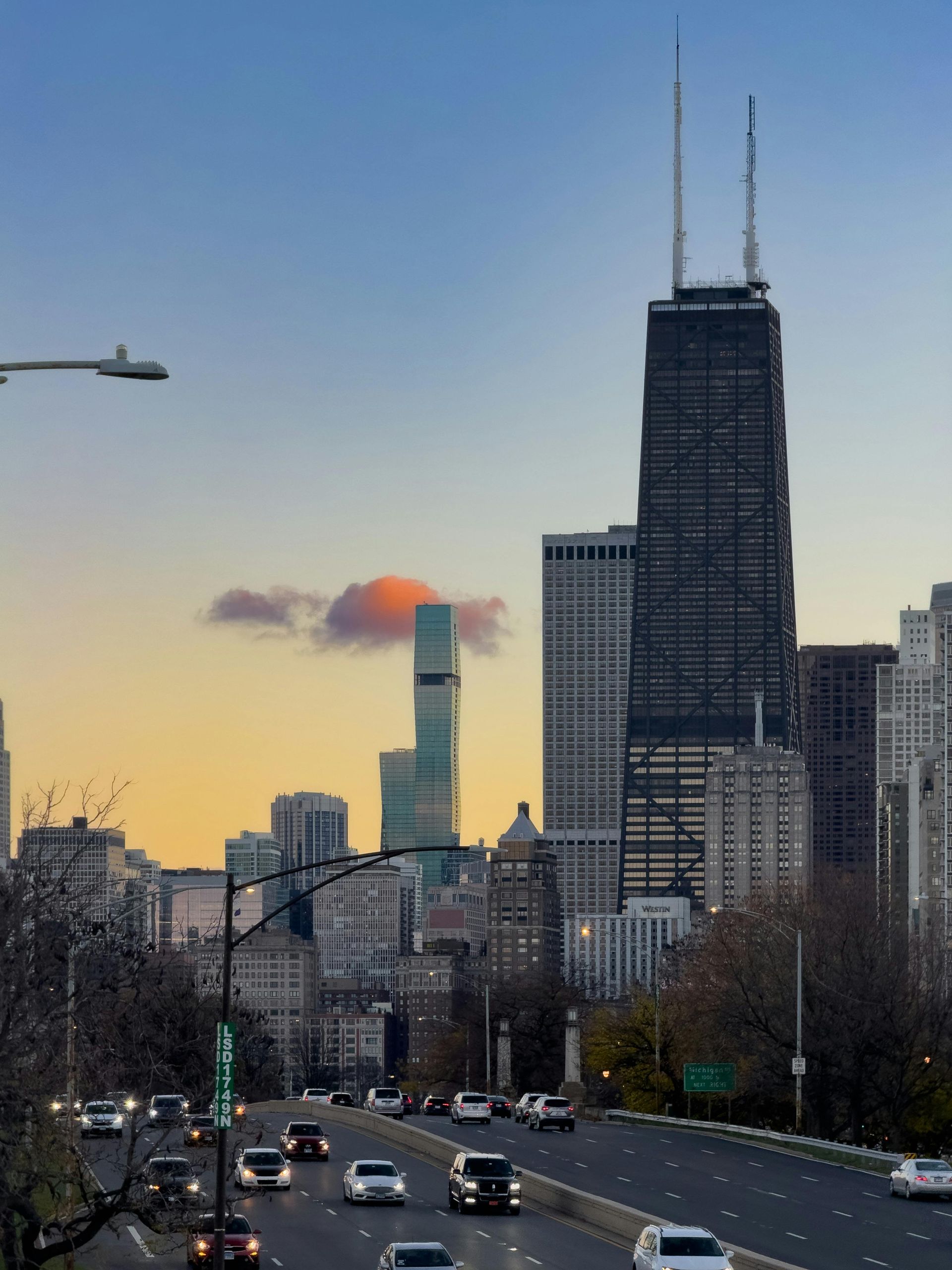City skyline at sunset with a tall black skyscraper and traffic on a busy road