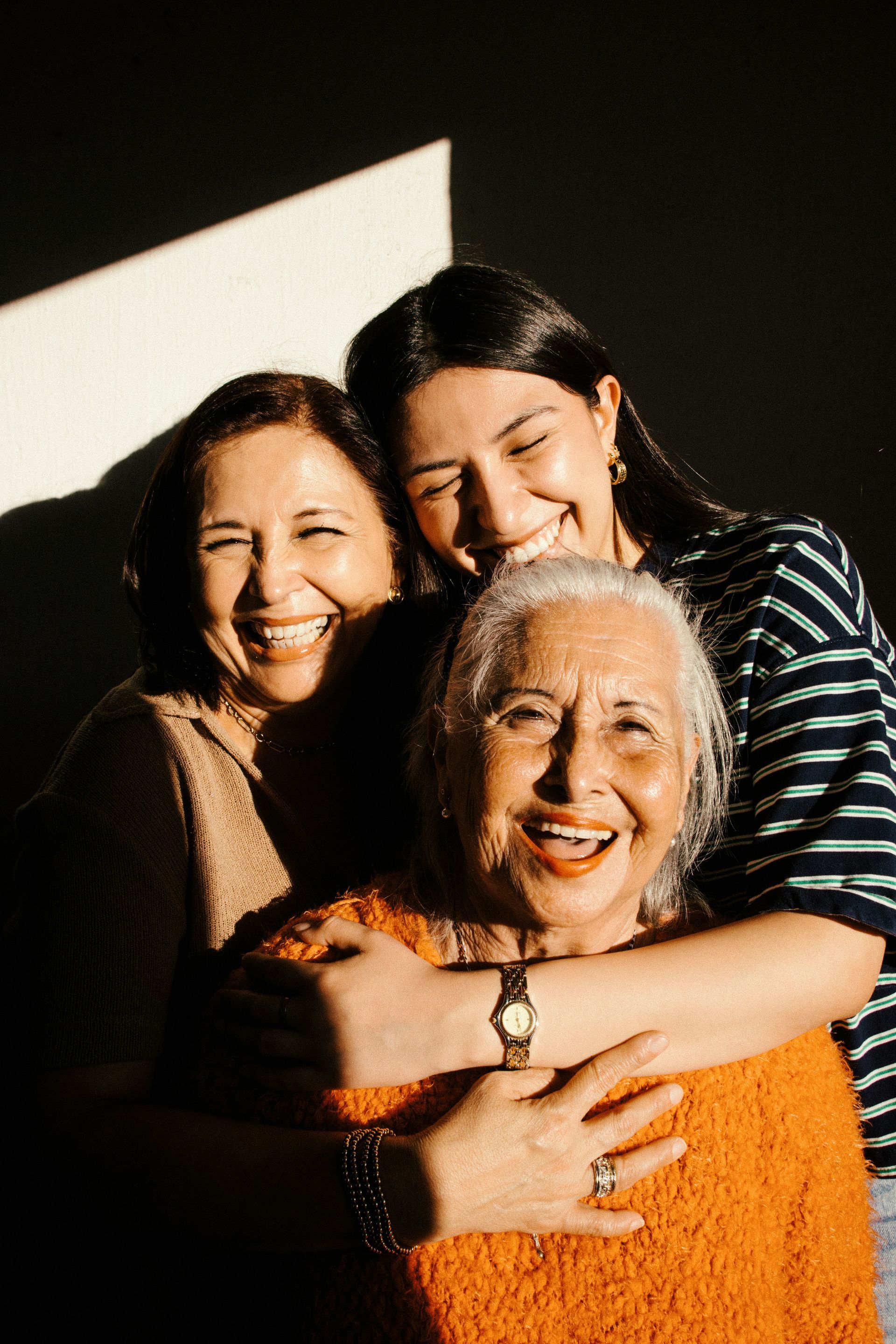 Three smiling people hugging in warm sunlight, with the front person wearing an orange sweater.