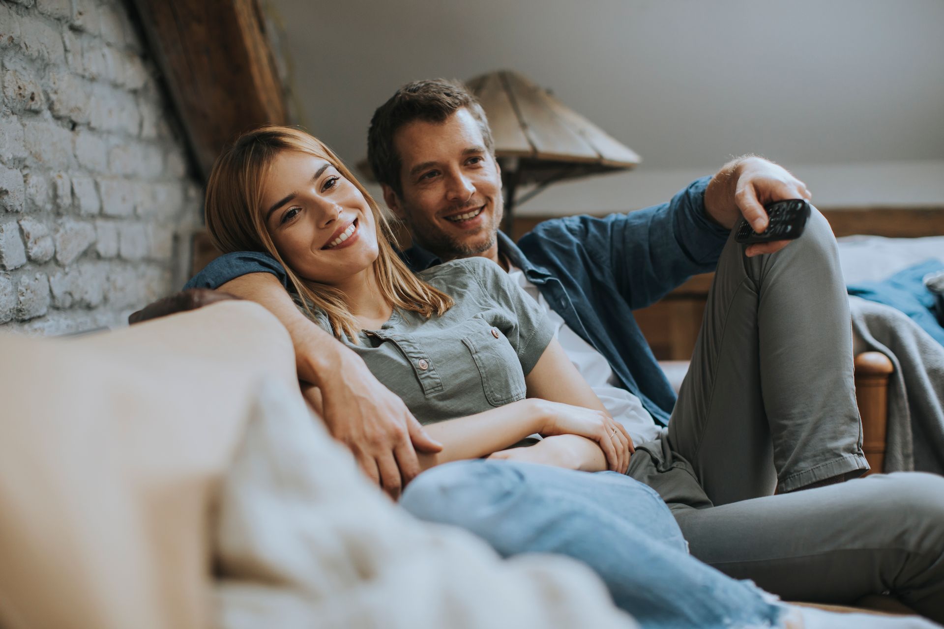 Two people relaxing on a couch, smiling in a cozy living room by a stone wall.