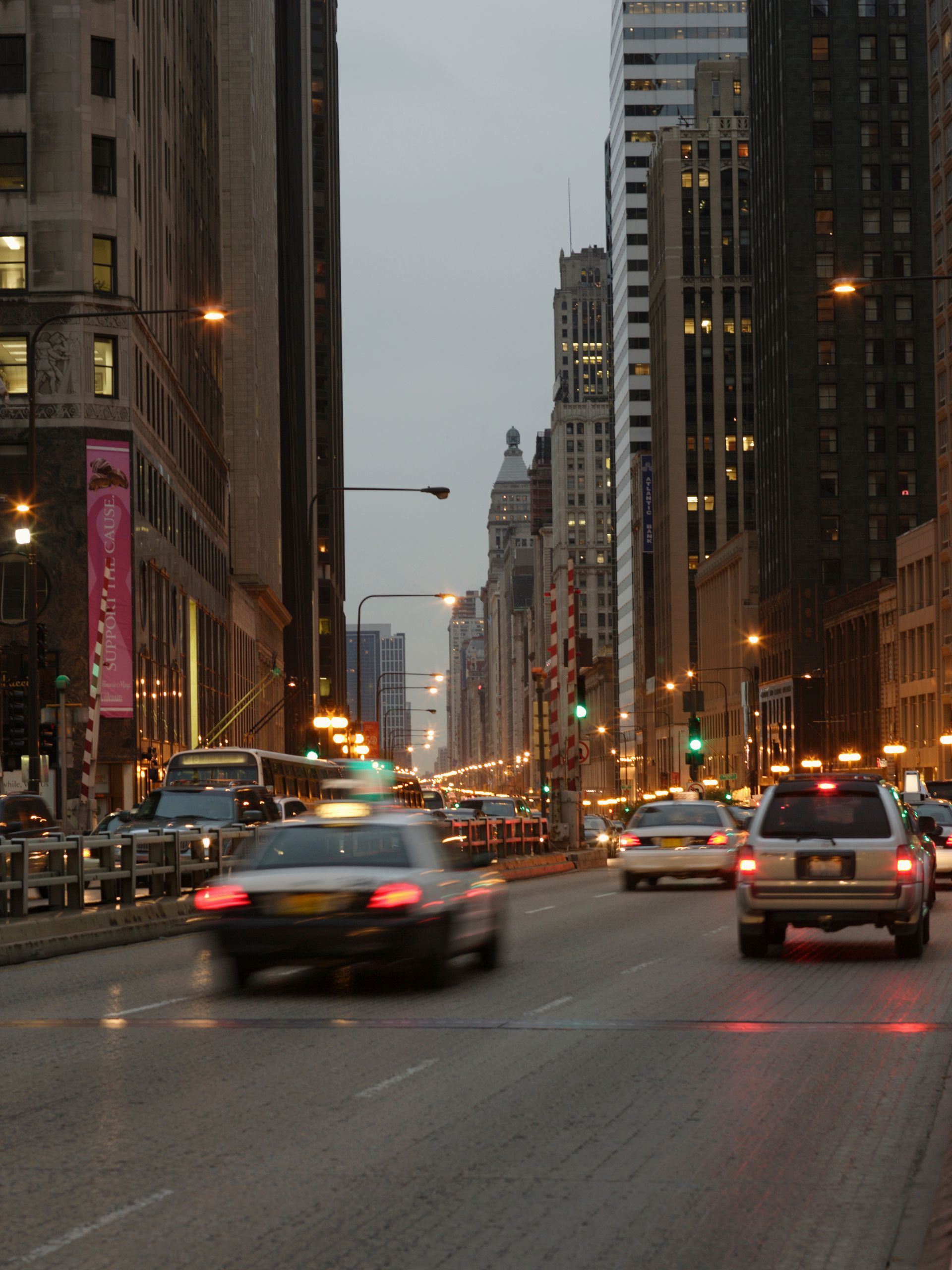 Cars driving on a wet city avenue at dusk between tall buildings and glowing streetlights