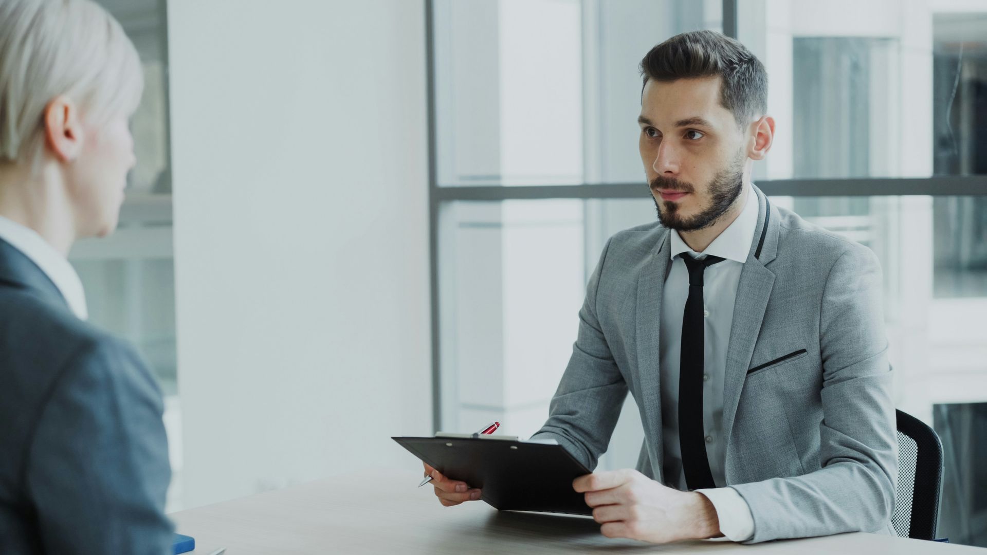 Two people in a bright office meeting, one holding a tablet while speaking across a desk.