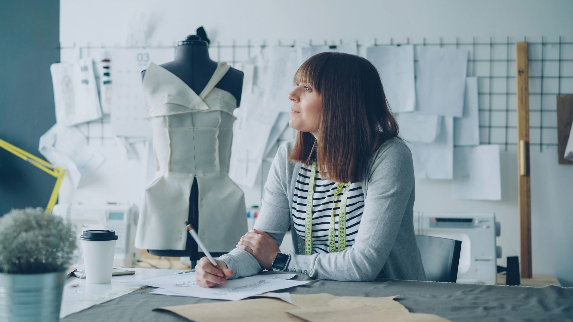 Person sketching a dress design in a sewing studio, with fabric patterns and dress forms nearby