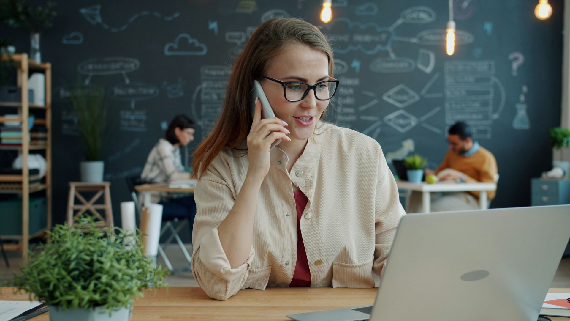 Woman working on a laptop in a café, wearing glasses and a beige shirt, with a chalkboard wall behind her.