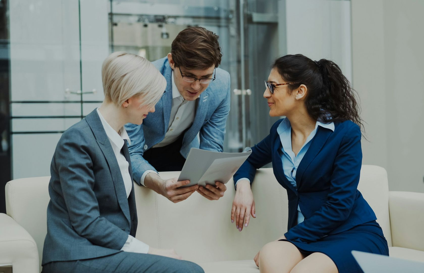 Three coworkers in business attire discussing a tablet in a modern office lounge