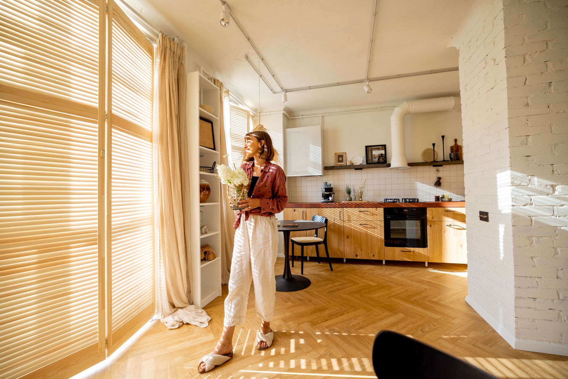 Person standing in a sunlit kitchen with wooden floors, holding a phone near large windows and blinds