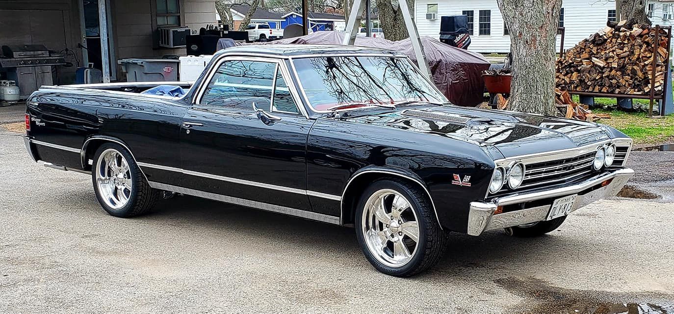 Black classic El Camino parked on a gravel driveway, chrome wheels, shiny black paint.