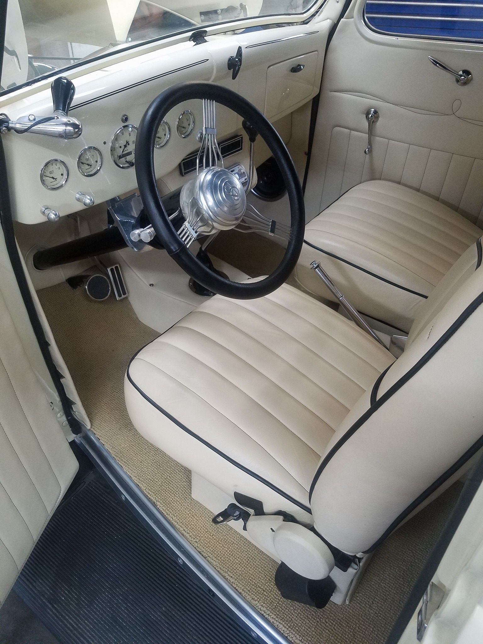 Cream-colored classic car interior with black steering wheel, gauges, and seats. Beige floor and door panels.
