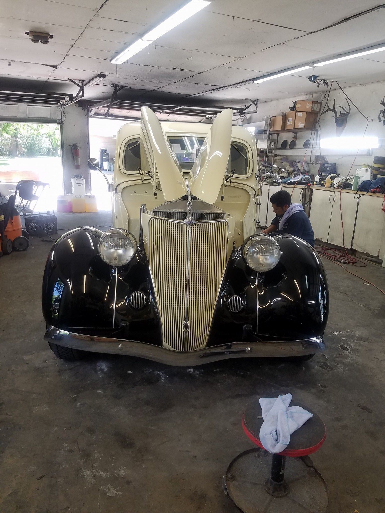 Vintage car in a garage with hood open, black body, cream top, person working.