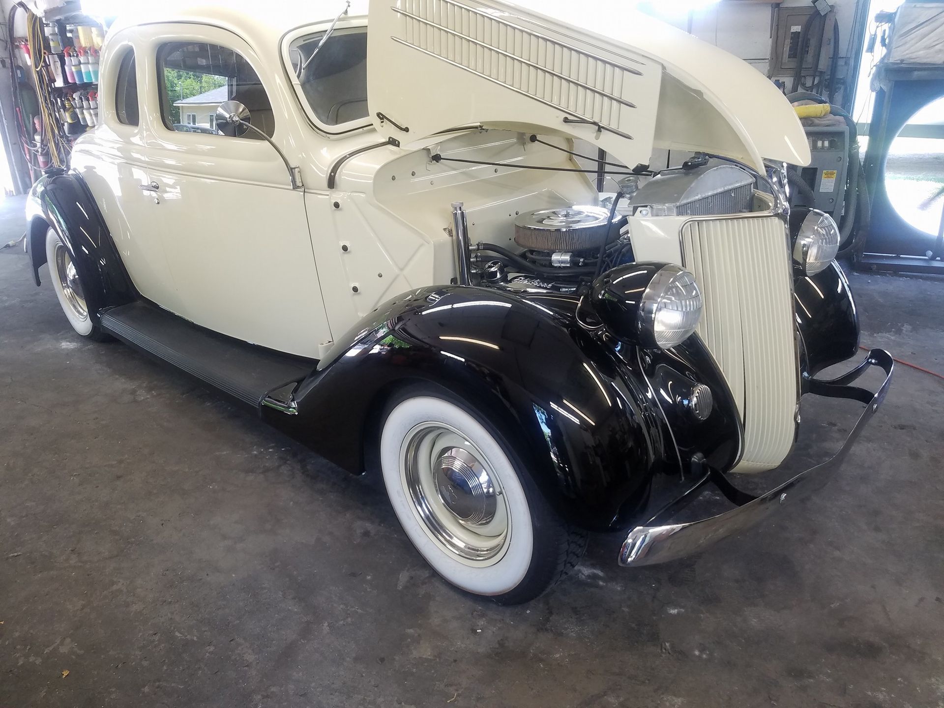 Cream and black classic car with hood open, in a garage.