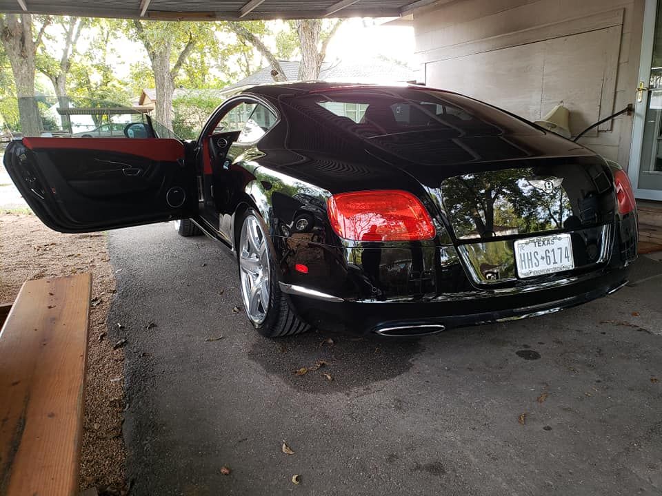 Black Bentley coupe with door open, parked under a shelter. Red interior visible.
