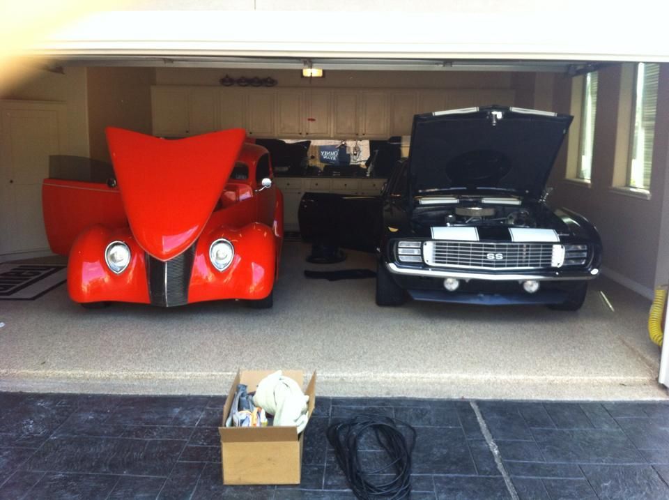 Two classic cars, one orange and one black, parked inside a garage with open hoods.