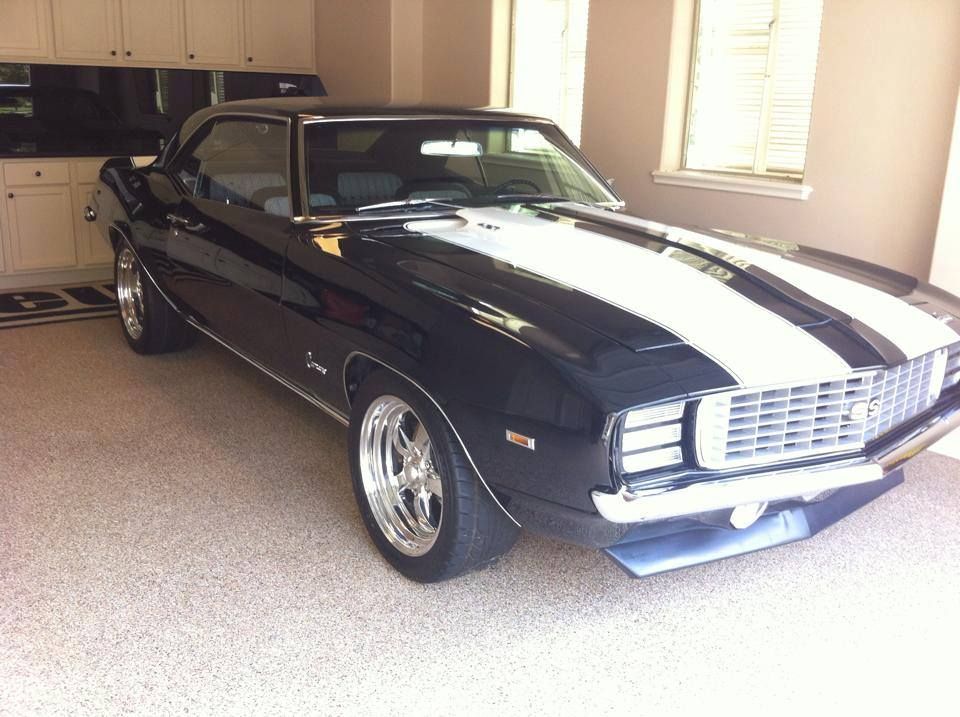 Black vintage Camaro with white racing stripes parked in a garage.